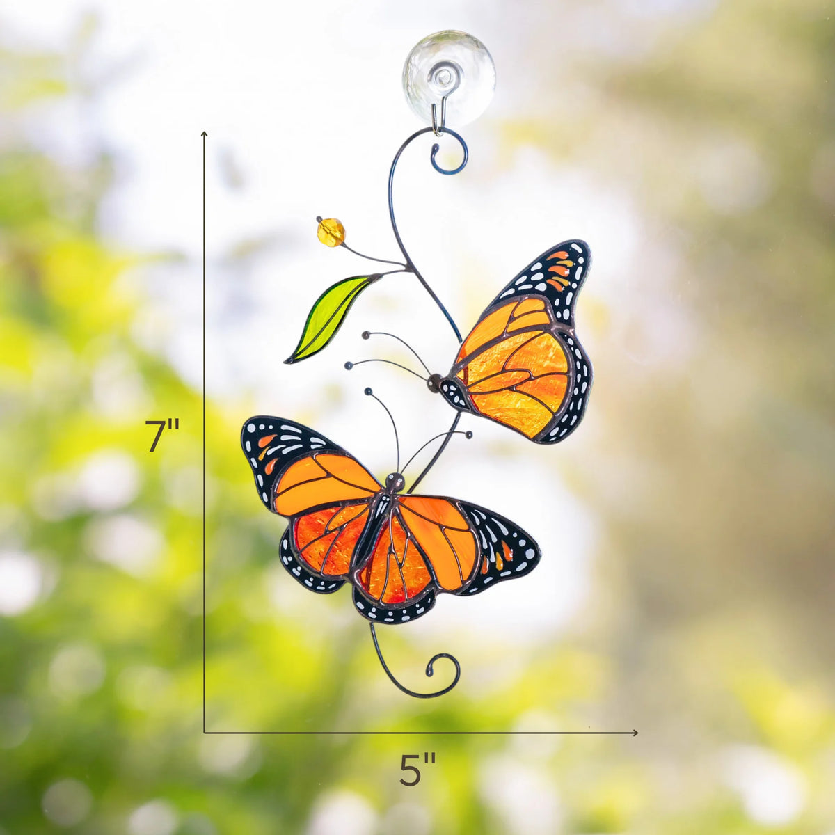 Monarch butterflies stained glass suncatcher measuring 7 by 5 inches, hanging from a clear suction cup with soft green garden bokeh behind the amber and orange wings