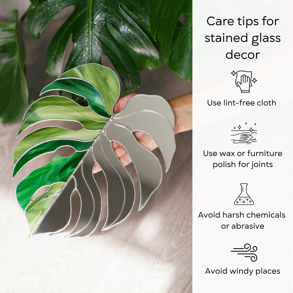 Hand holding the Monstera Leaf Stained Glass Mirror under dewy monstera leaves - care for this handmade stained glass decor with a lint-free cloth, wax the solder joints, avoid harsh chemicals