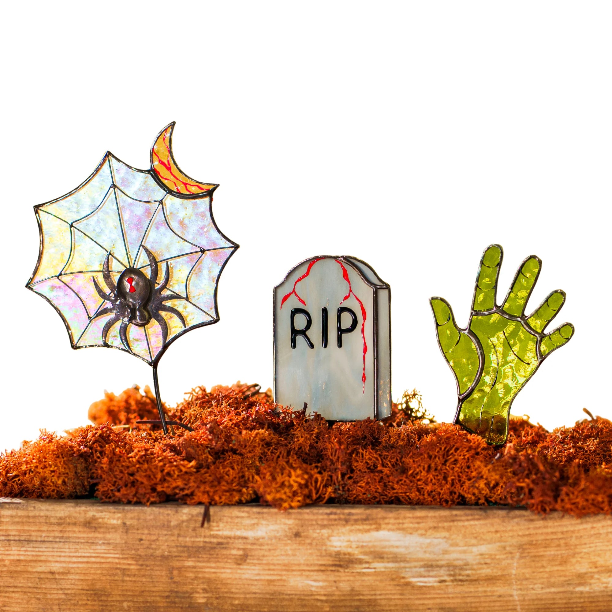 Stained glass tombstone, zombie hand, and spiderweb figures nestled in bright orange moss on a wooden surface, ideal for holiday gifting.