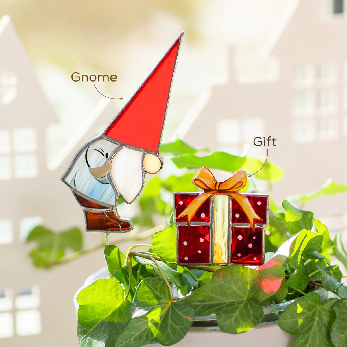 Christmas gnome in a red pointed hat and a red gift box with a gold bow - two handmade stained glass plant stakes in a pot of ivy, festive Christmas decor for houseplants
