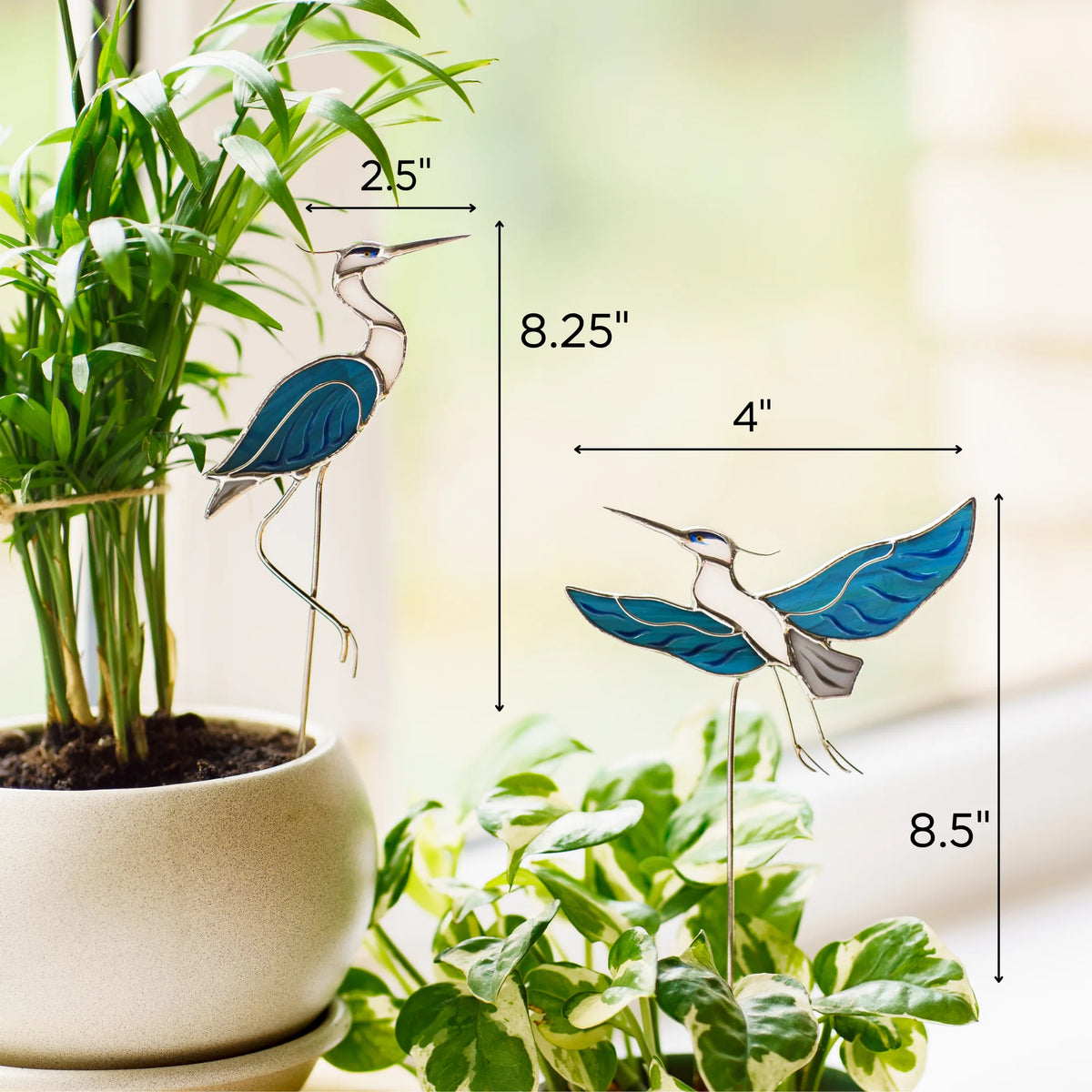 Size chart for stained glass heron plant stakes - sitting heron 2.5 by 8.25 inches, flying heron 4 by 8.5 inches - shown beside houseplants on a sunlit windowsill
