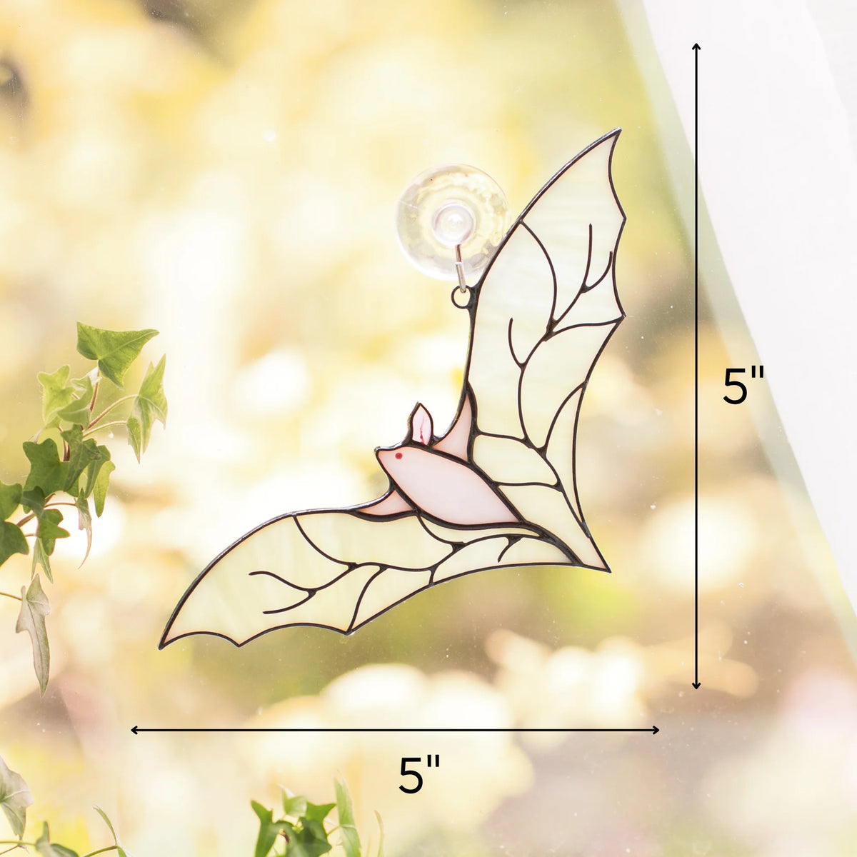 White stained glass bat suncatcher measuring 5 by 5 inches, hanging by suction cup on a sunlit window with green plants nearby.