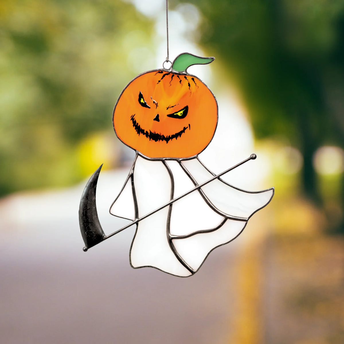 Two hands cradling the grim reaper pumpkin head stained glass suncatcher with golden autumn leaves scattered in the background.