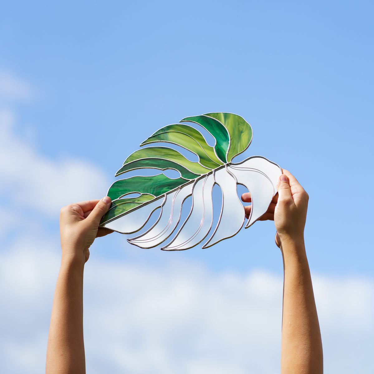 Hands holding the Monstera Leaf Stained Glass Mirror up to a blue sky, the mirror half reflecting soft clouds beside swirled green glass with leaf cutouts - handmade botanical stained glass art