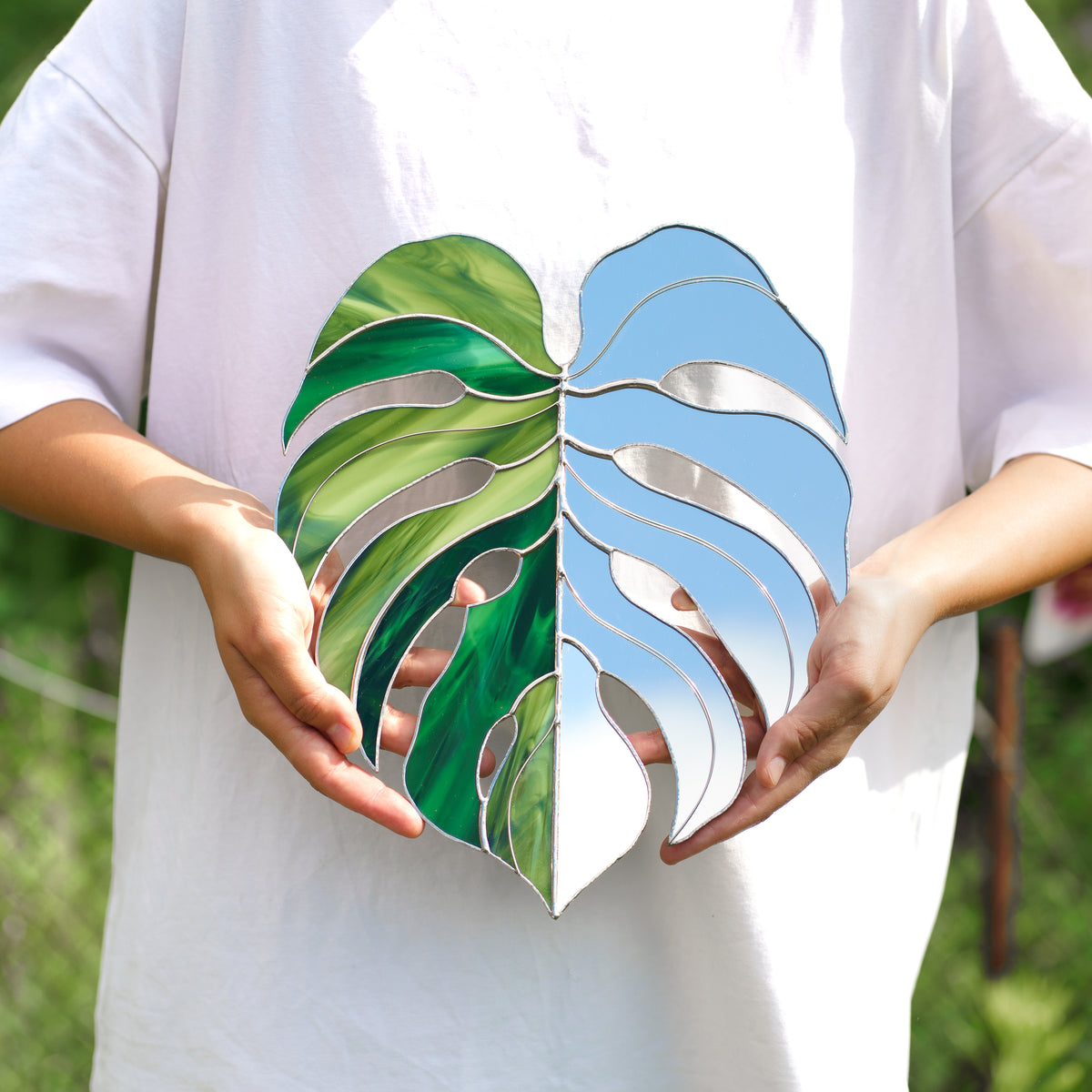 Person in a white t-shirt holding a handmade Monstera Leaf Stained Glass Mirror, the mirror half reflecting blue sky next to emerald and lime green glass - a unique housewarming gift