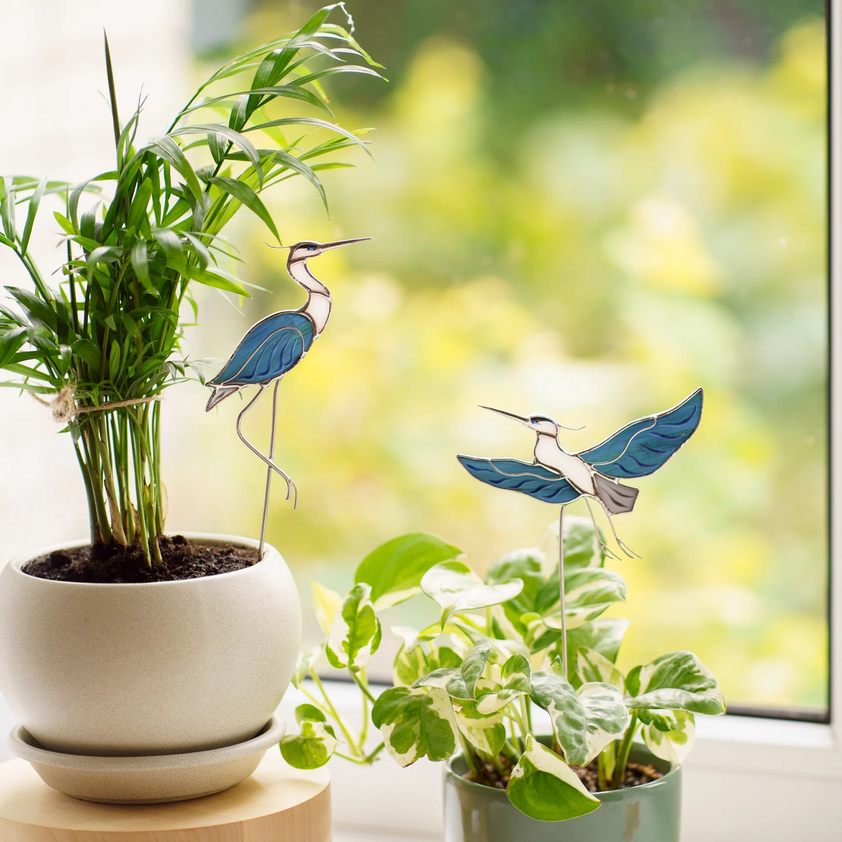 Stained glass sitting and flying heron plant stakes in blue glass displayed in potted houseplants on a bright windowsill, handmade garden decor for bird lovers