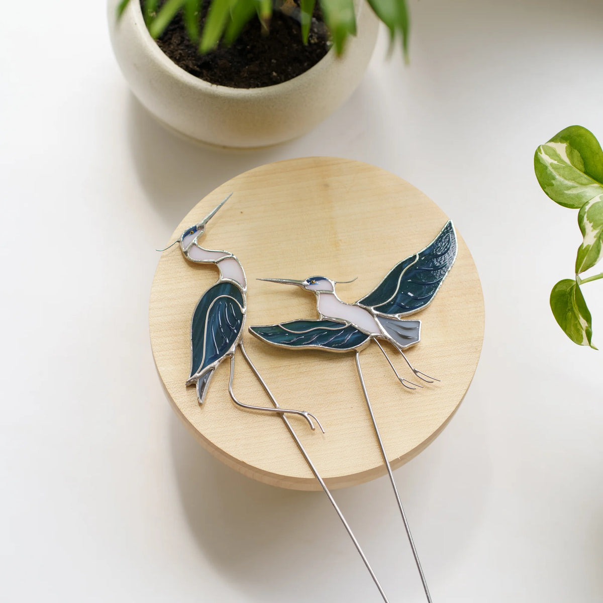 Top-down view of two stained glass heron plant stakes - sitting and flying poses in blue, white and grey glass - resting on a wooden stand beside a potted plant