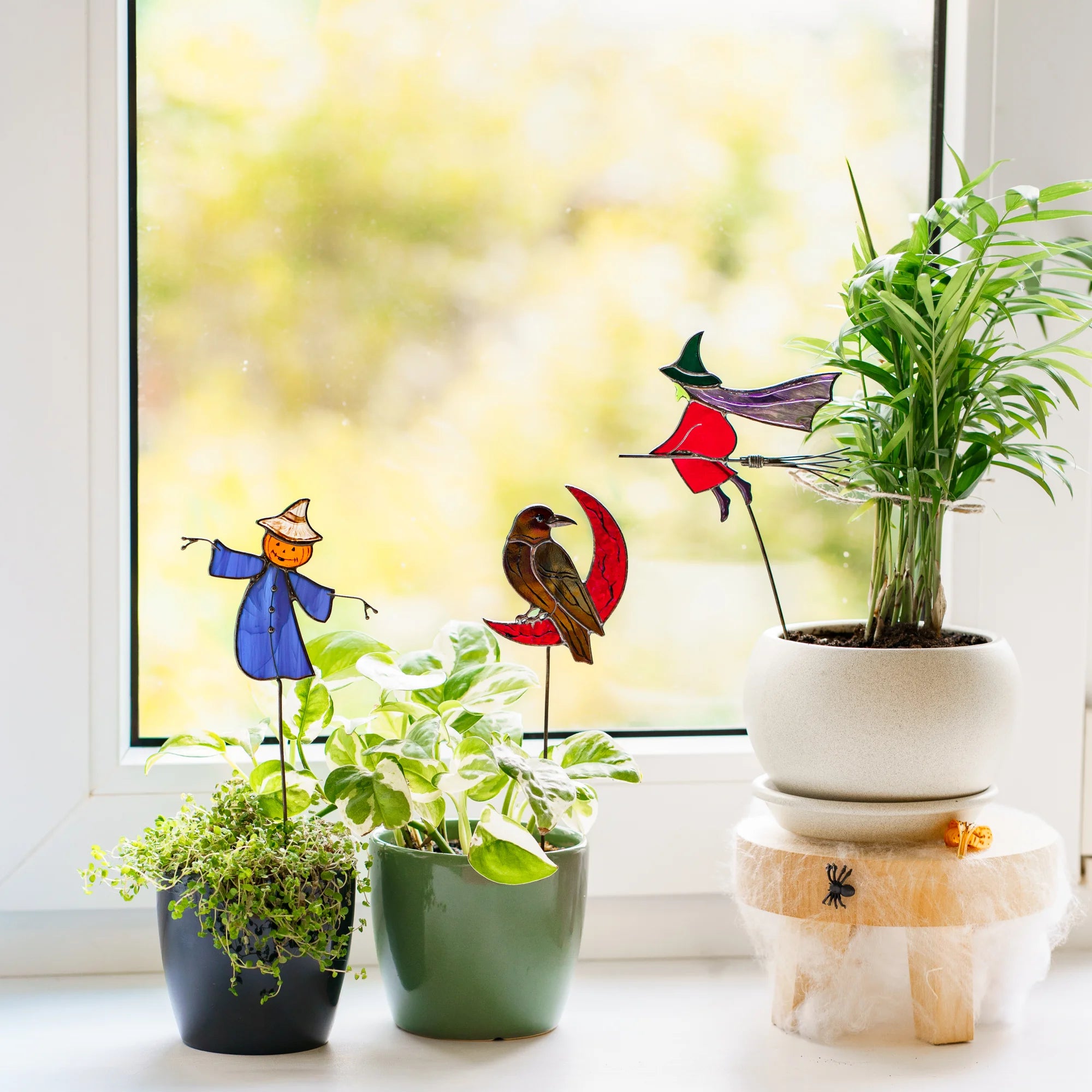 Halloween stained glass plant stakes on a sunlit windowsill - a pumpkin-headed scarecrow in a blue coat, a raven on a red crescent moon and a witch on a broom among potted plants
