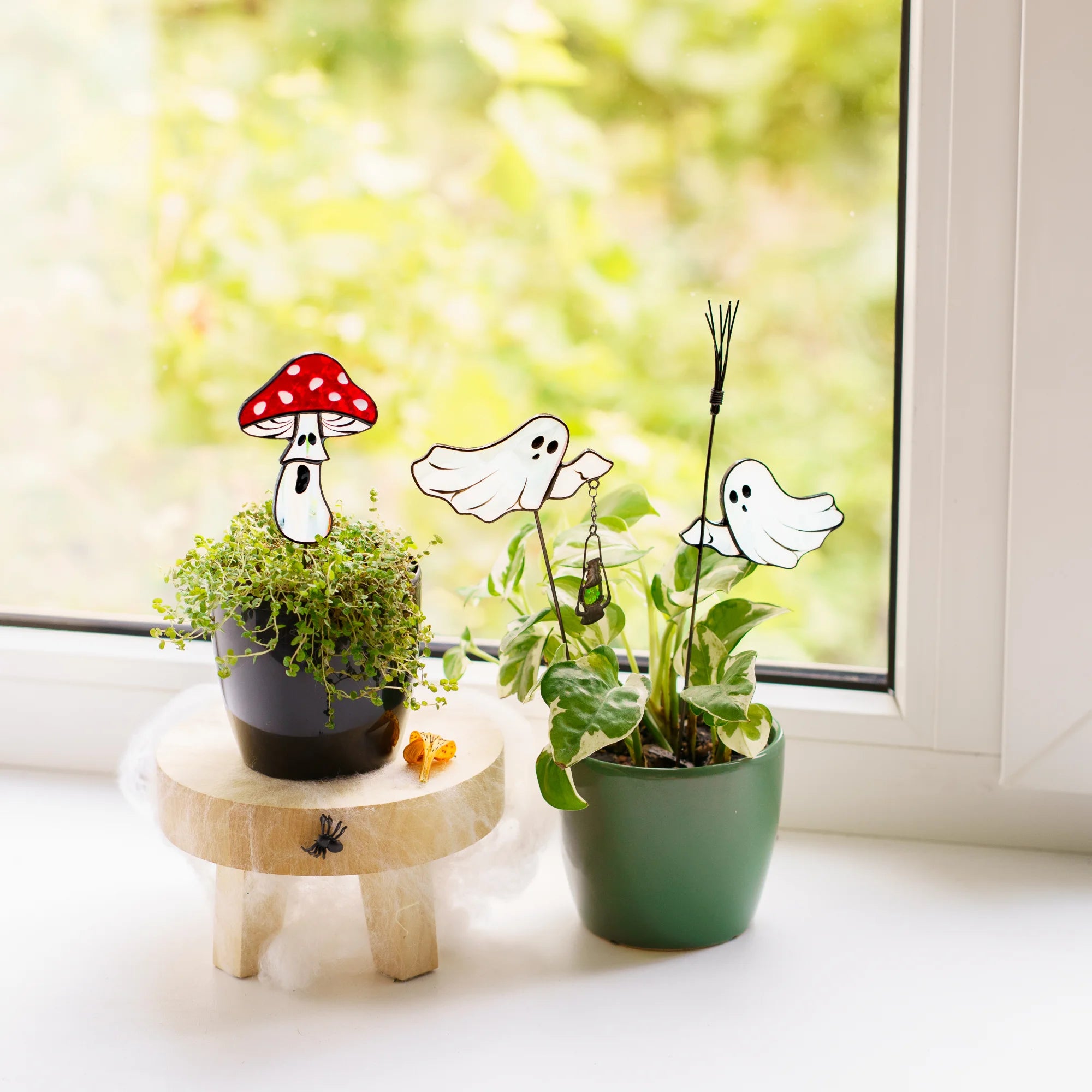 Fly agaric mushroom stained glass plant stake with a startled face beside two glass ghosts tucked into a pothos pot, handmade Halloween garden art glowing in soft window light