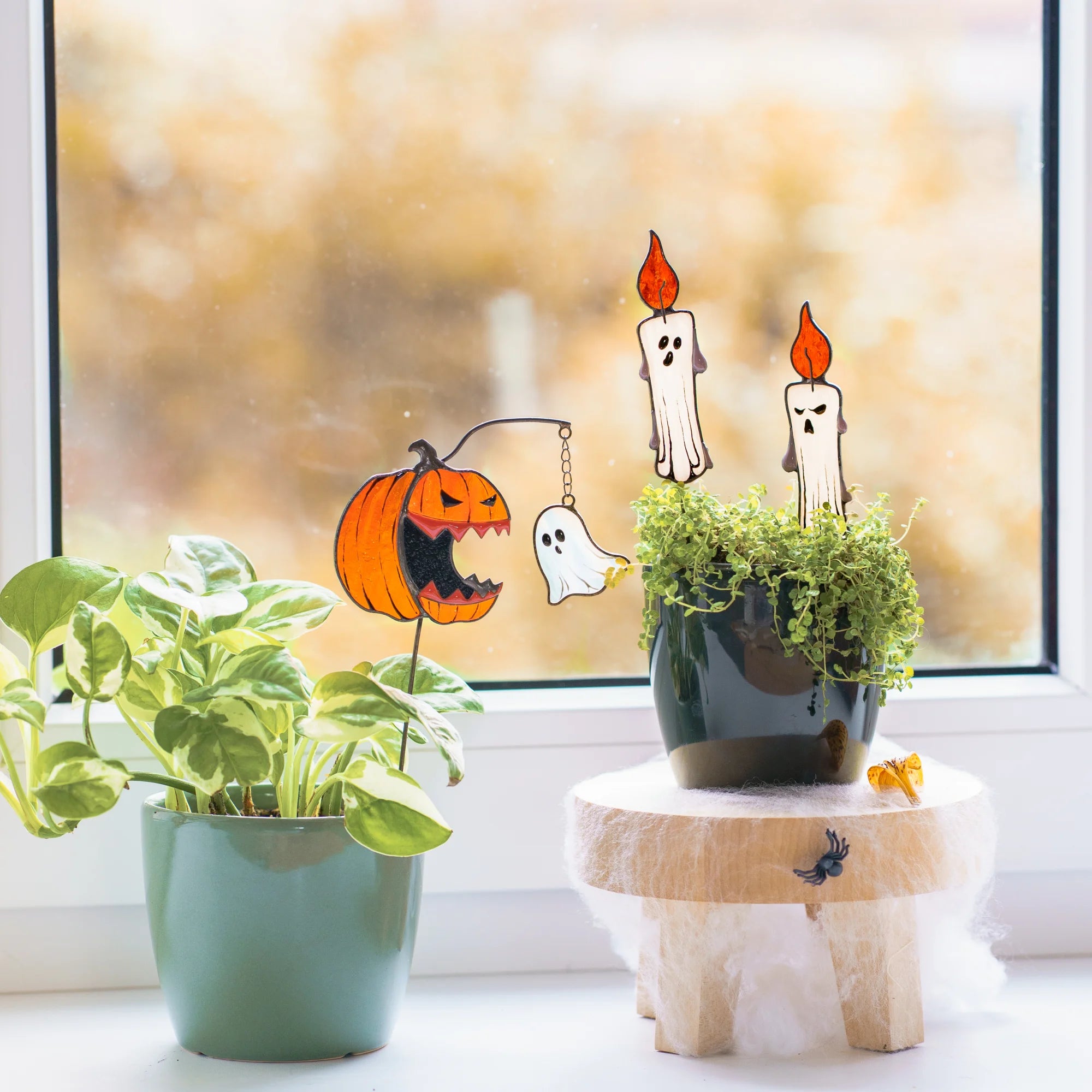 Halloween-themed window stained glass plant stakes displayed on a window sill next to a green potted plant. The set includes a jack-o'-lantern with a hanging string, a ghost, and two candle cutouts with flames.
