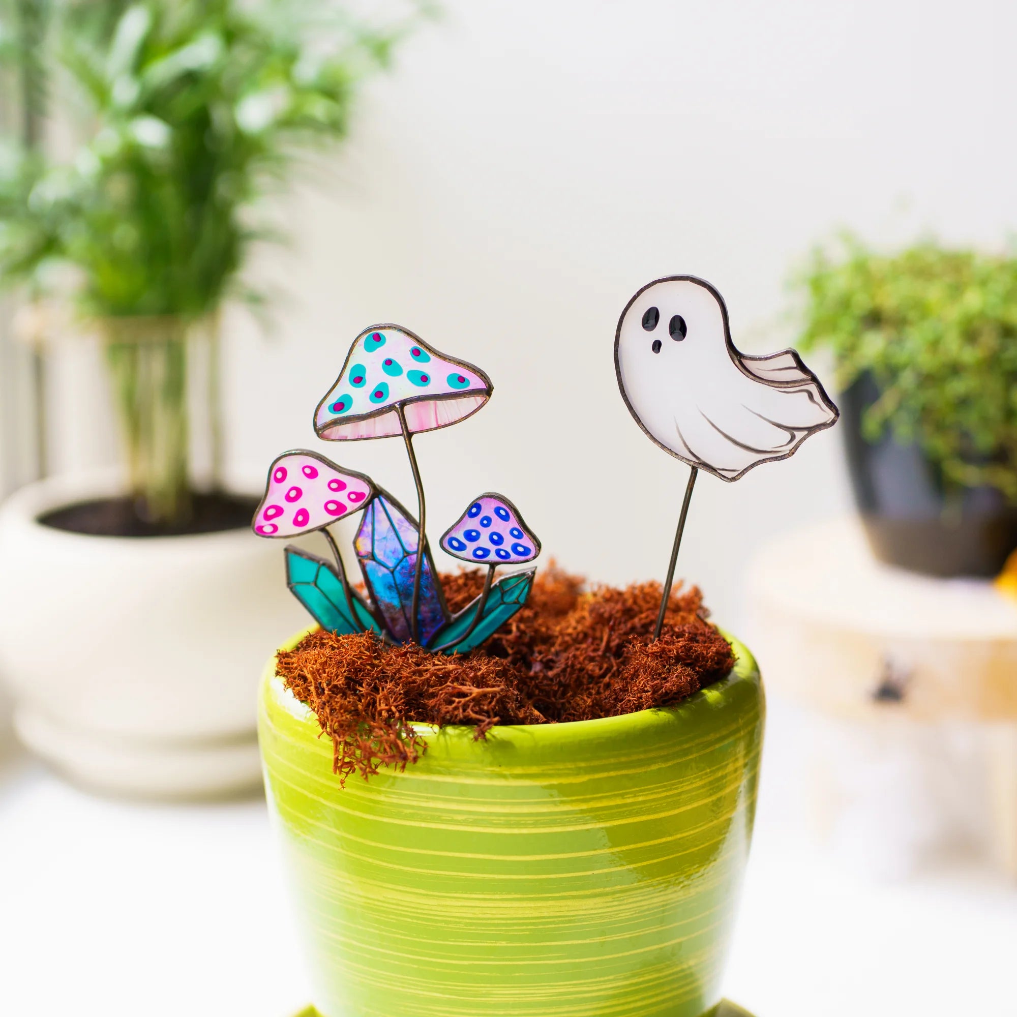 Stained glass mushroom and ghost plant stakes in a lime green pot of moss - three iridescent pink toadstools around a blue crystal beside a small white ghost with black eyes