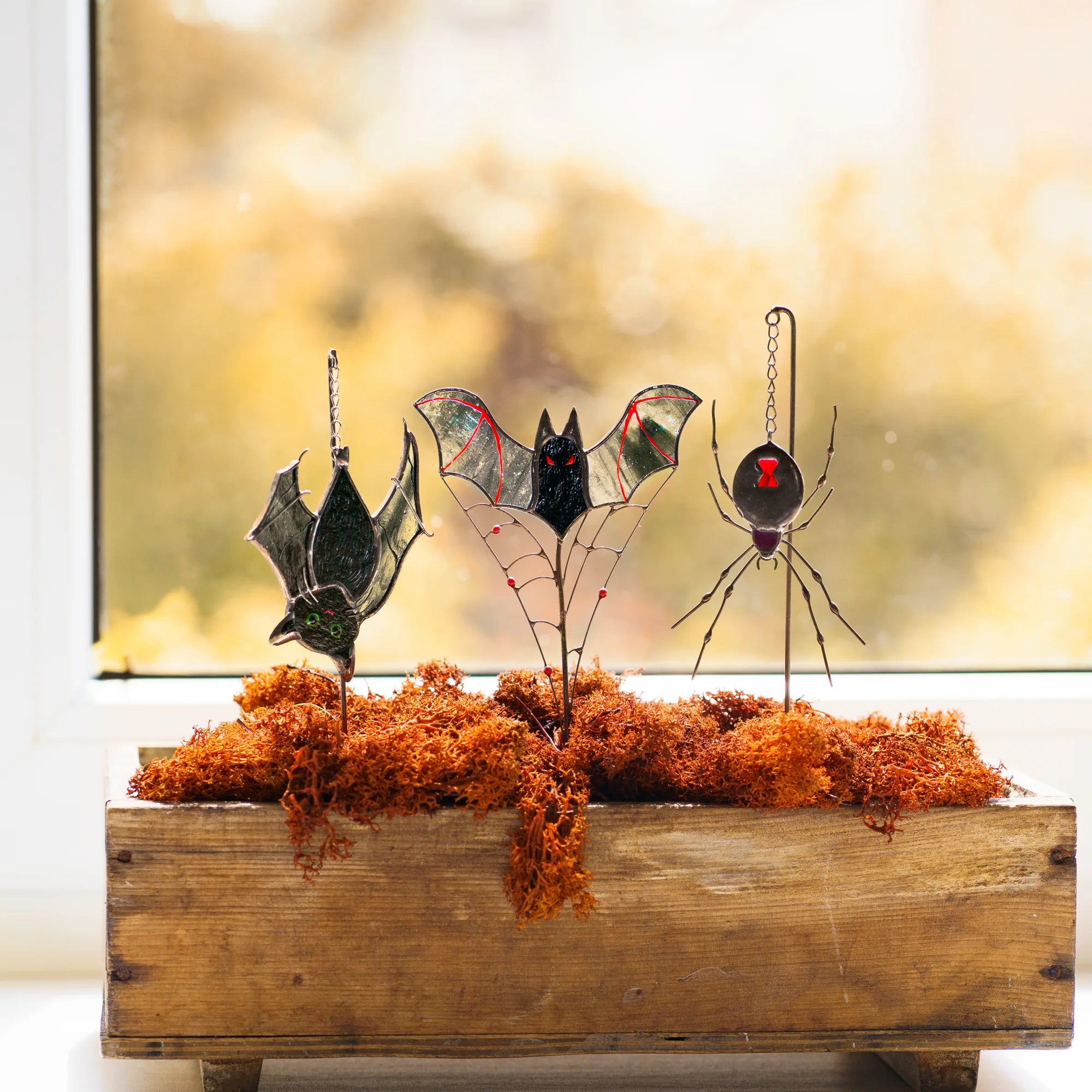 Halloween stained glass plant stakes in a wooden box of rust-orange moss on a sunlit windowsill - a red-eyed vampire bat on a wire web, a black widow spider and a cat-bat on chains