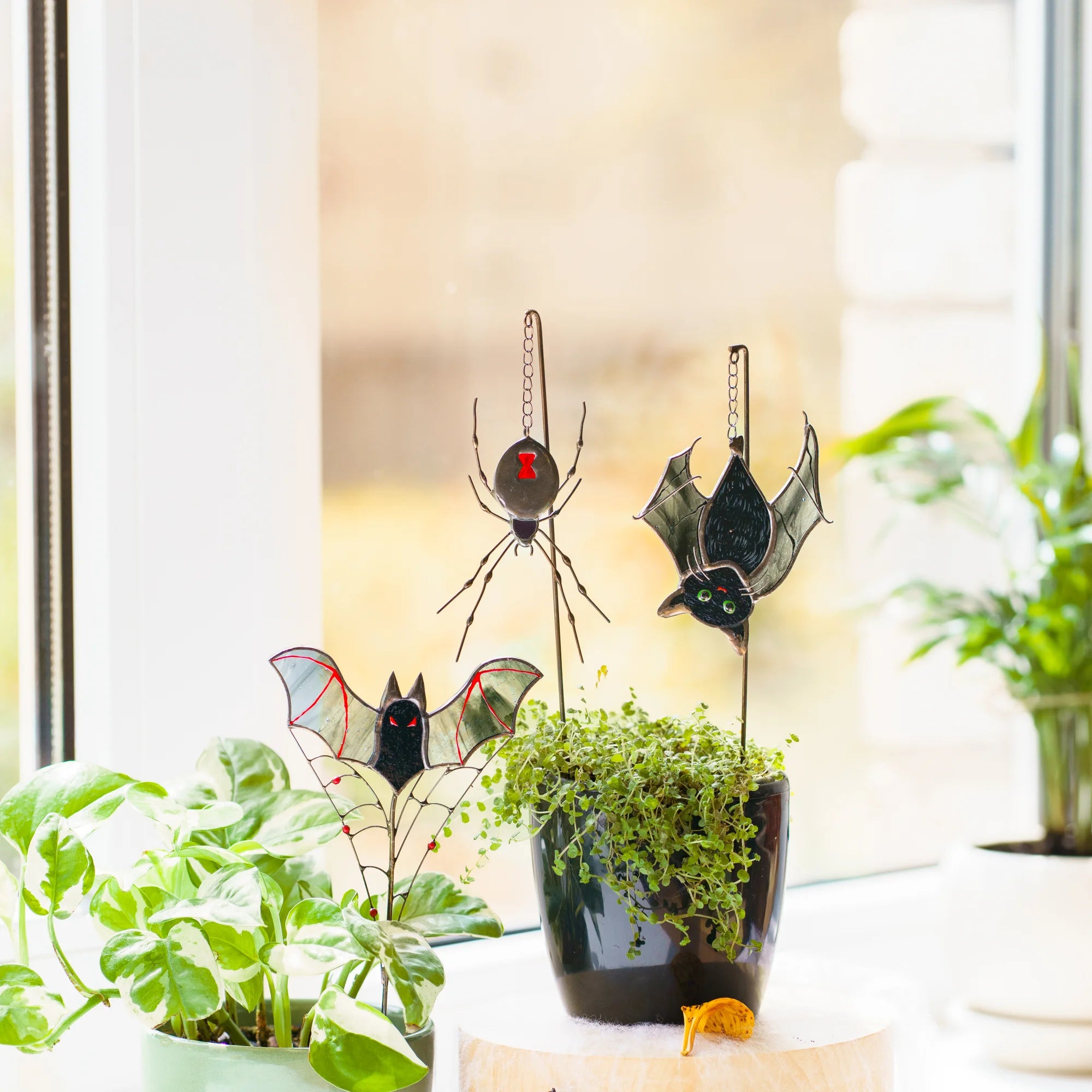 Three handmade bat and spider plant stakes among houseplants - stained glass black widow with a scarlet hourglass, a cat-bat with green eyes and a grey-winged bat on a spiderweb