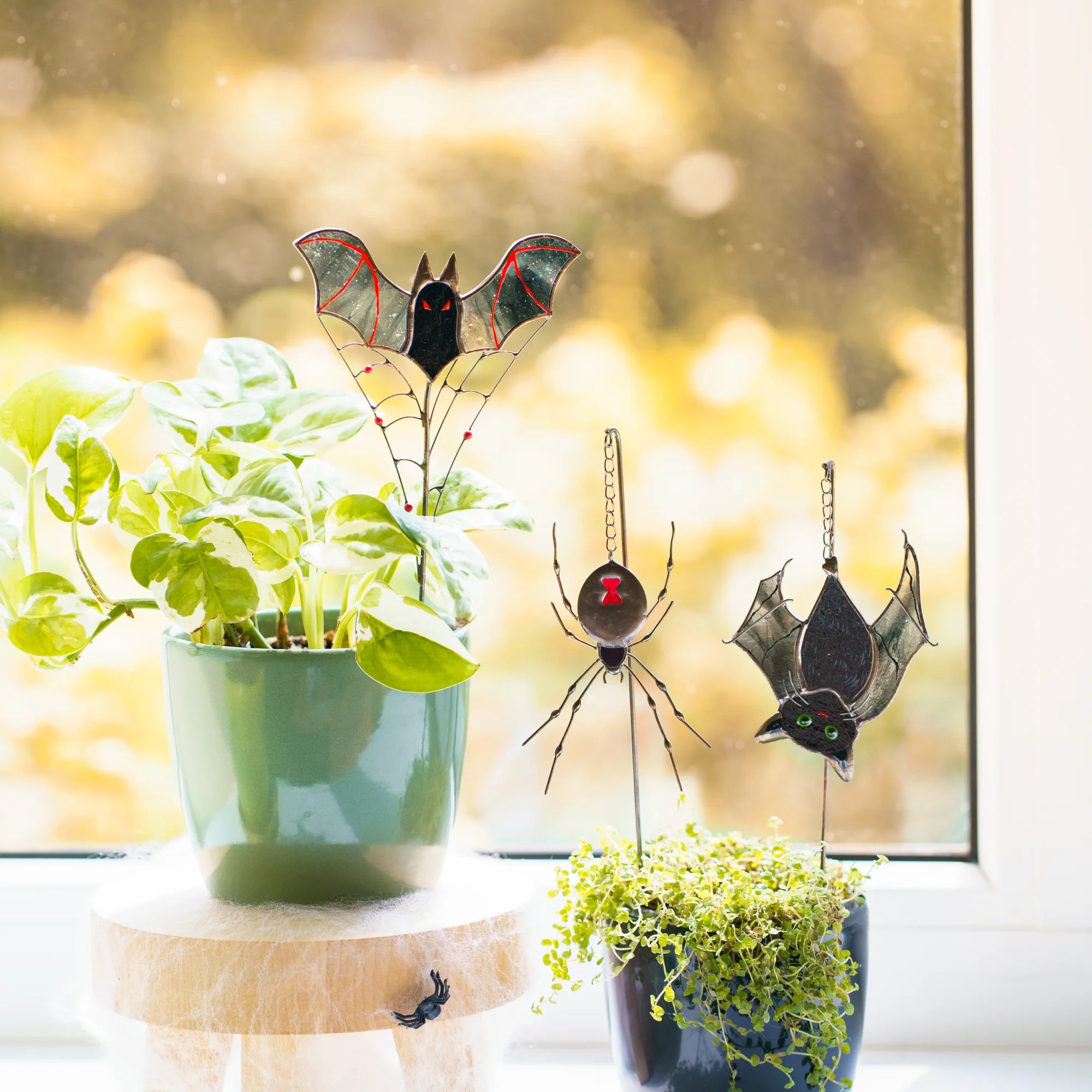 Stained glass bat plant stake with smoky grey wings and red veins rising above a pothos, beside a black widow spider and a hanging cat-bat - spooky window decor for plant lovers