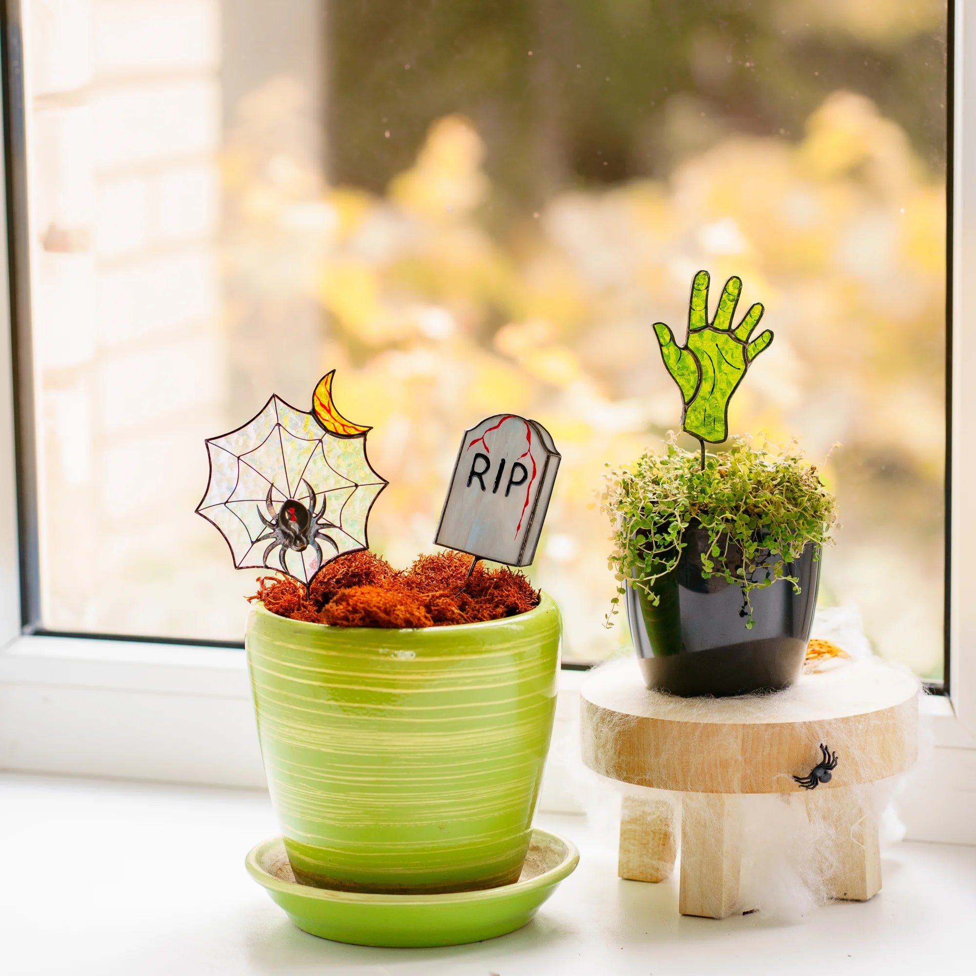 A spooky stained glass Halloween plant stake set featuring a spiderweb with a spider and moon, a RIP tombstone, and a green zombie hand, making a wonderful holiday gift.
