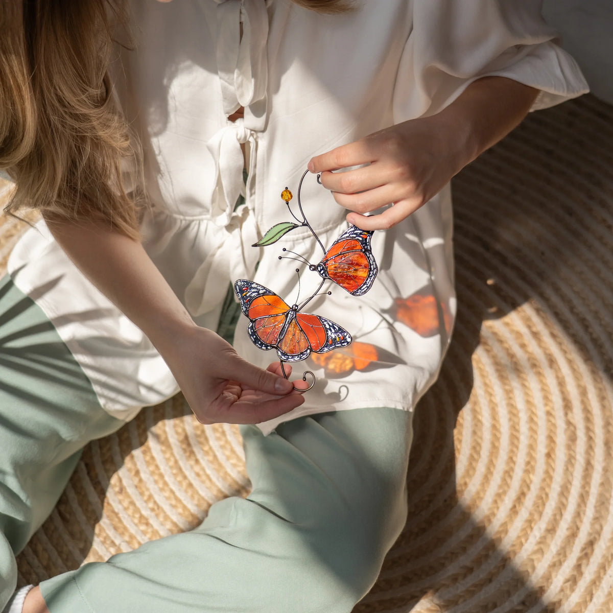 Monarch butterfly stained glass suncatcher held in both hands above a jute rug, its wings throwing warm amber patches of light across a cream blouse - handmade window decor