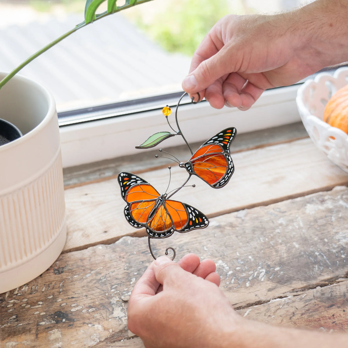 Hands holding a stained glass pair of monarch butterflies over a weathered wooden windowsill beside a white ribbed planter - handmade orange wings, wire stem and green leaf