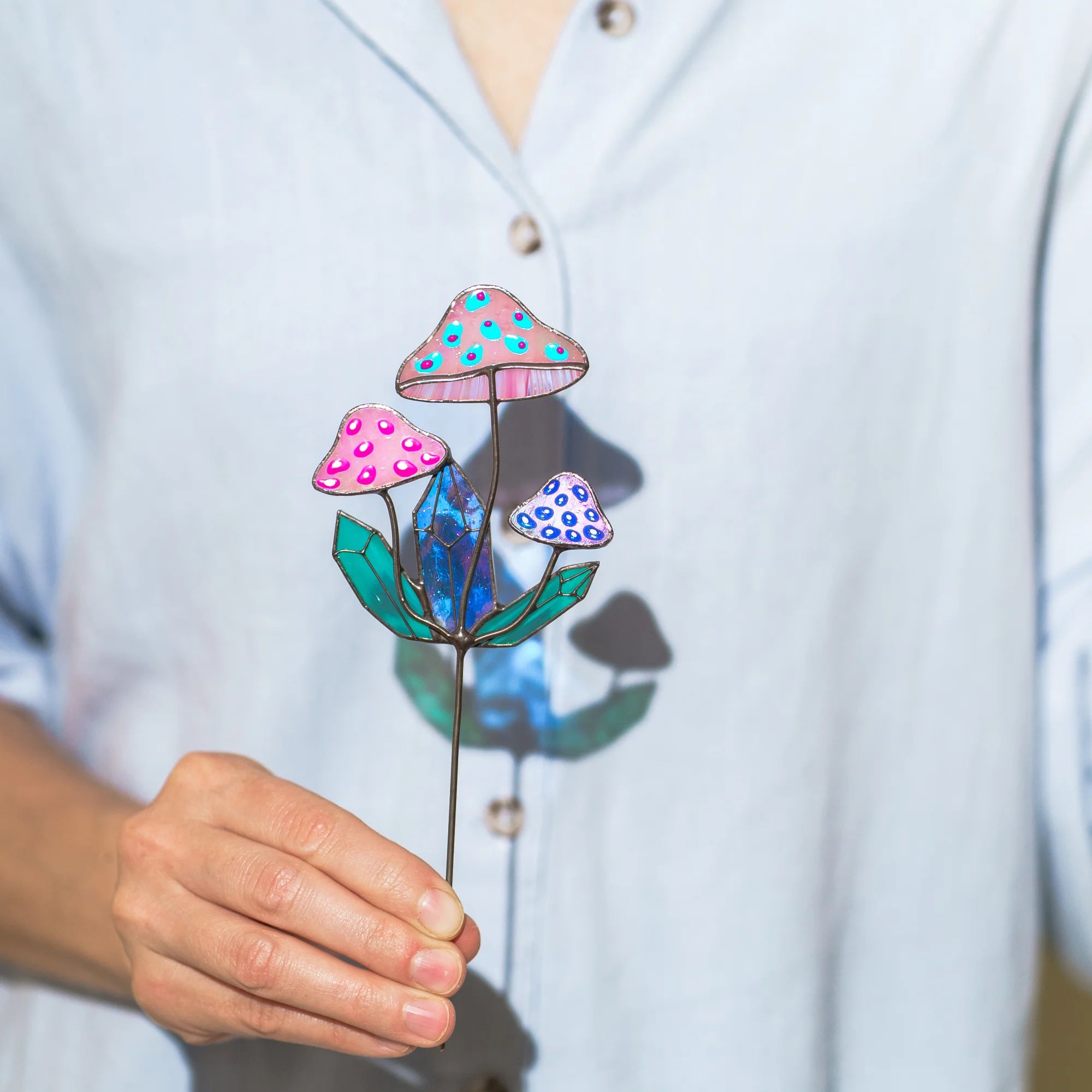 Woman holding an iridescent stained glass mushroom plant stake - three spotted pink toadstools and teal crystal shards around a blue-violet crystal, on a slim metal stake