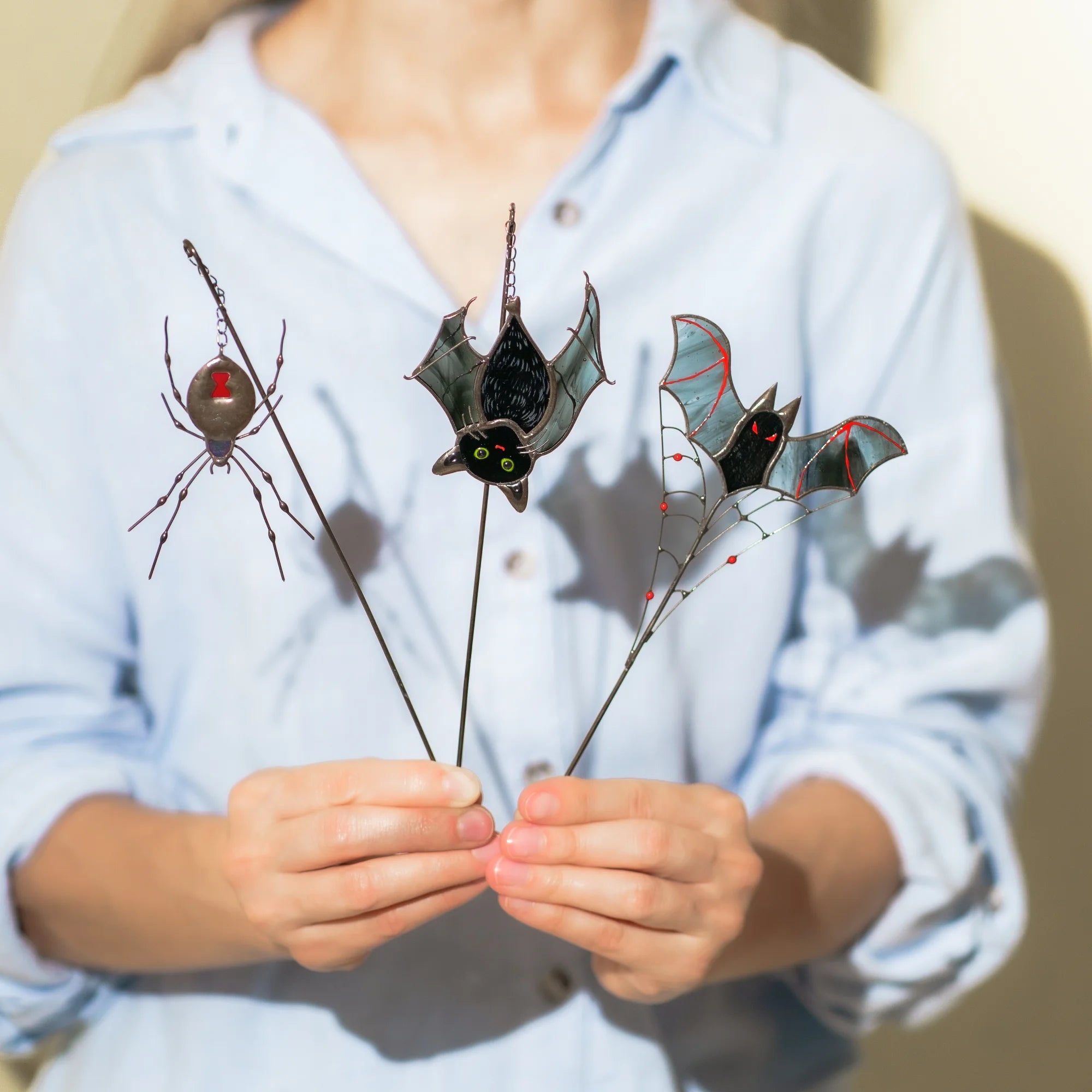 Woman holding three handmade stained glass plant stakes - black widow spider on a chain, black cat-bat with green eyes and a red-eyed vampire bat perched on a wire spiderweb