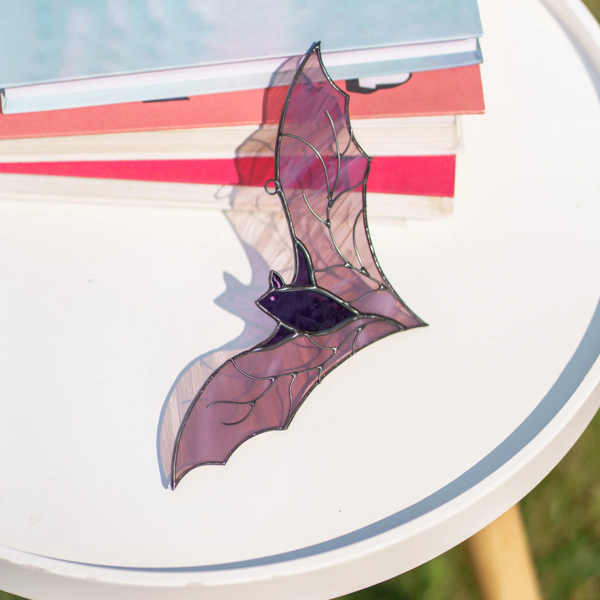 Handmade purple bat stained glass suncatcher lying on a white table beside a stack of books, sunlight casting a soft pink shadow of its wings across the surface