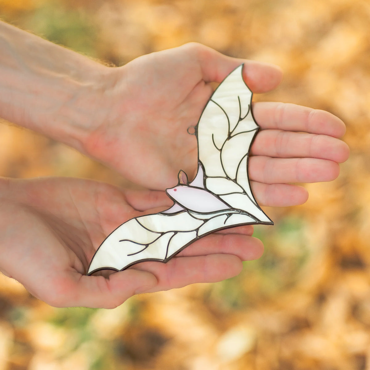 Hand cradling the white stained glass bat suncatcher in an open palm, set against a softly blurred golden autumn leaf background.