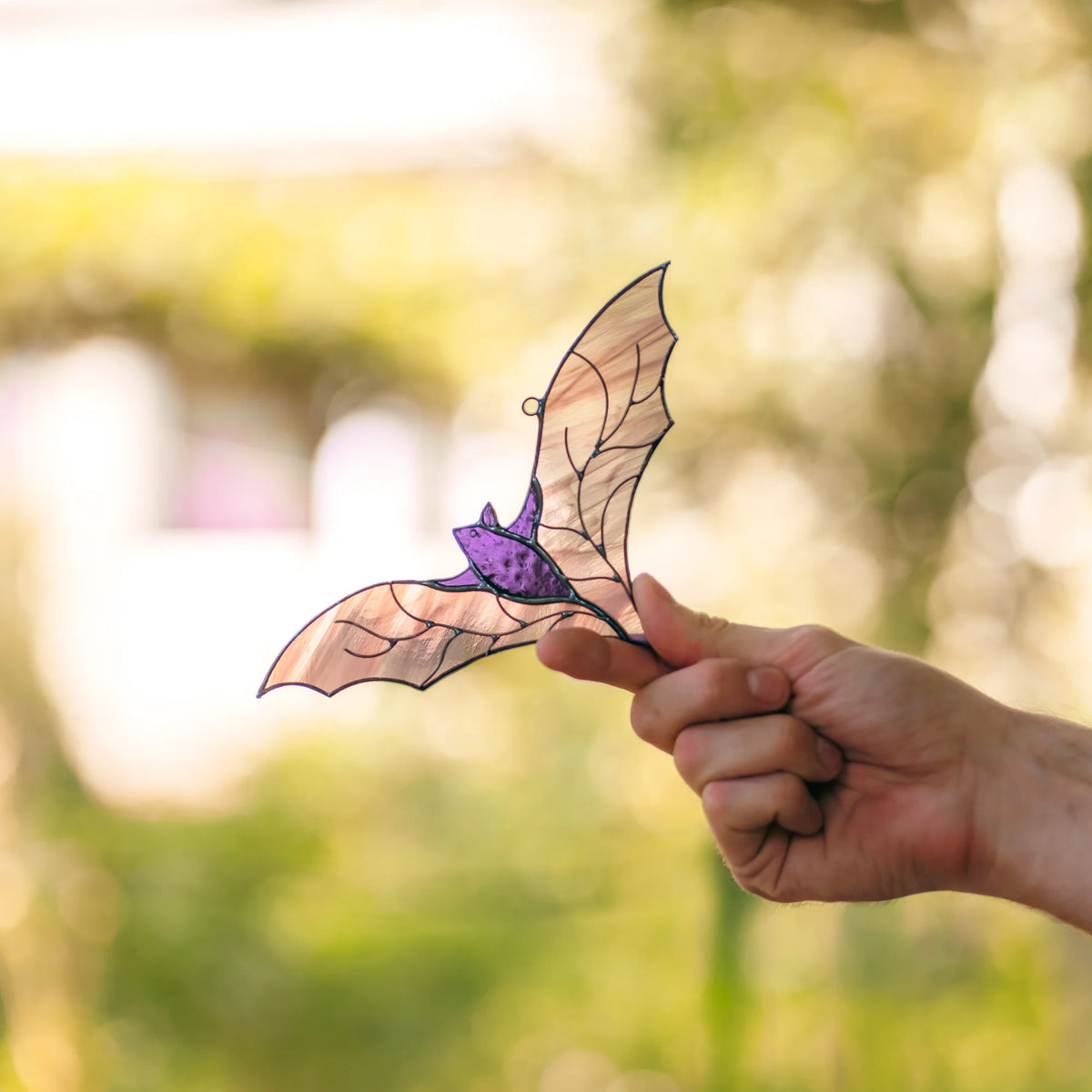 Hand holding the handmade purple bat stained glass suncatcher against a green garden background, showing the iridescent wings and dark purple textured body