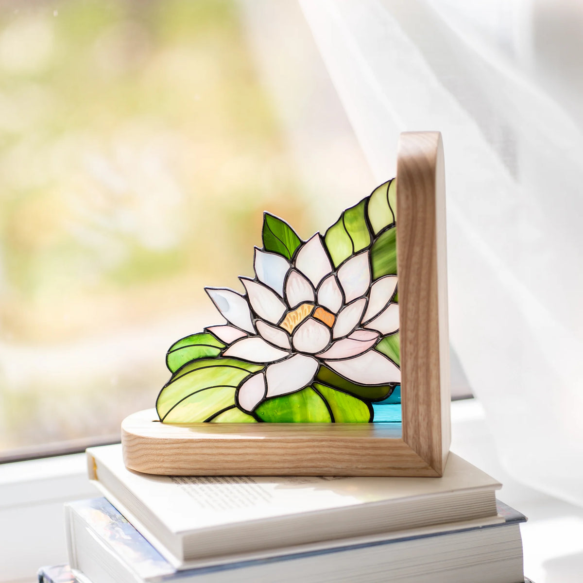 Stained glass water lily bookend resting on a stack of books beside a sunlit window, light ash wood frame glowing warm in natural daylight