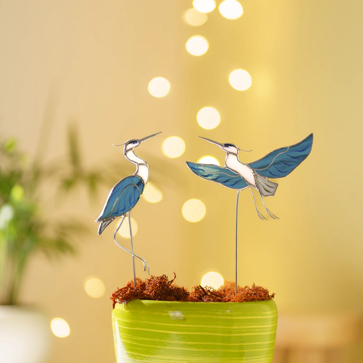 Stained glass heron plant stakes in a moss-topped pot with warm string lights in the background, a cozy handmade gift for bird lovers and plant lovers