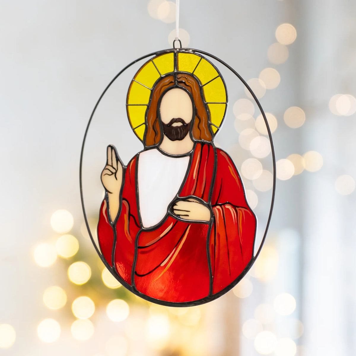 Jesus stained glass suncatcher hanging against glowing bokeh lights, the yellow halo and red glass robe shining in a slim oval frame - handmade hanging stained glass and a meaningful faith gift