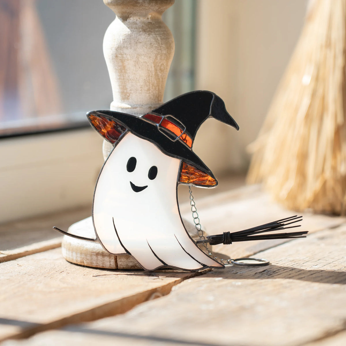 Stained glass ghost in a witch's hat resting against a wooden candlestick base on a plank surface, with a broom blurred in the background, handmade Halloween suncatcher decor