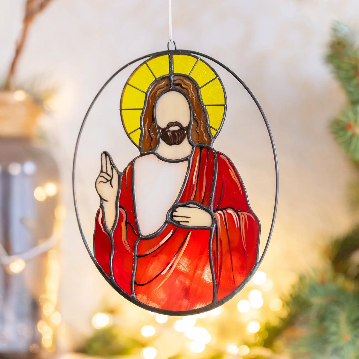 Jesus stained glass suncatcher hanging near pine branches and warm string lights, painted folds on the red robe catching the glow - handmade Christmas stained glass for windows and holiday decor
