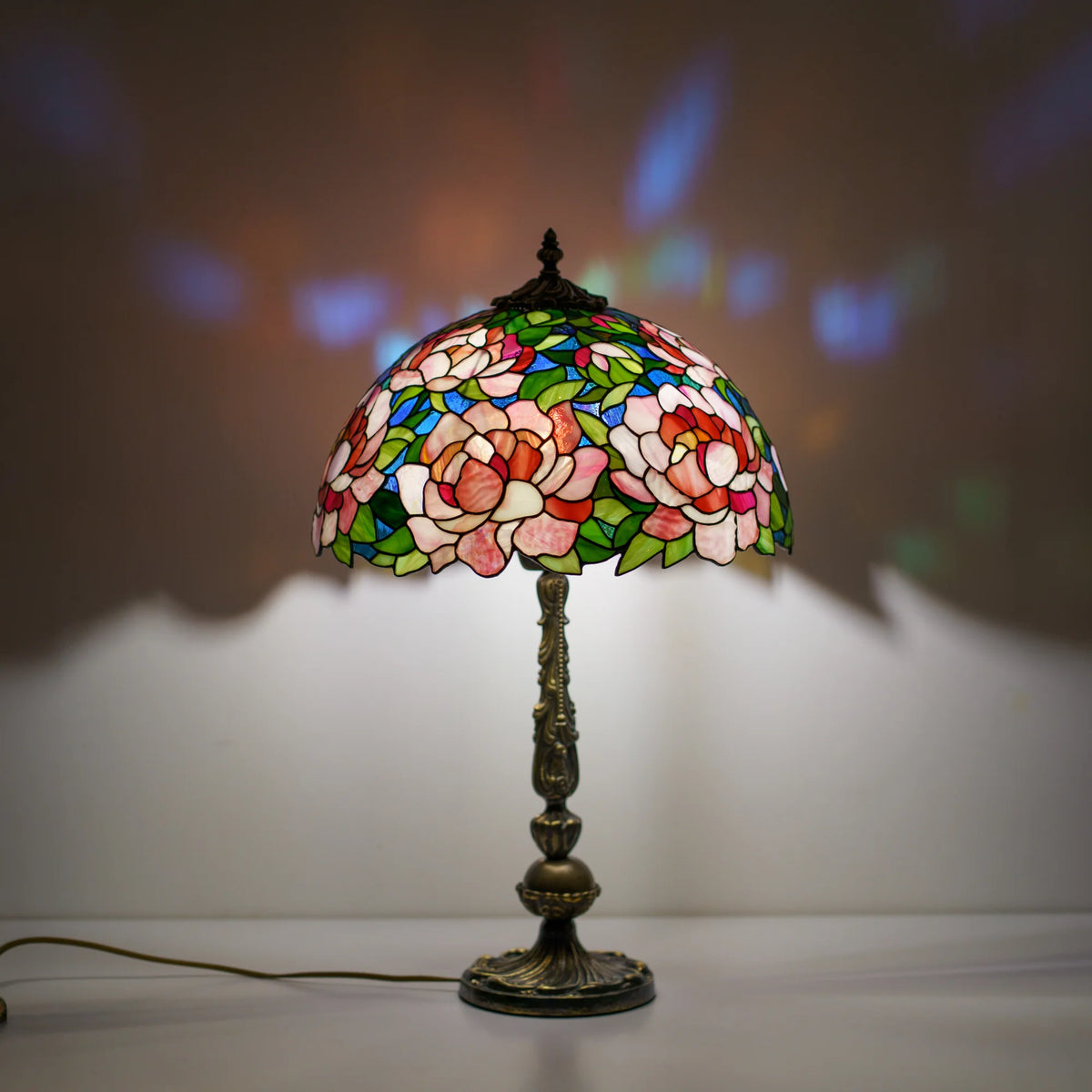 Handmade stained glass peony table lamp lit on a white pedestal, colorful stained glass light reflections glowing behind it in a dark room - a Tiffany style lamp for cozy evening ambiance