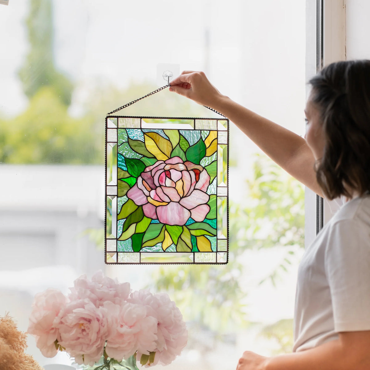Woman's hand hanging a stained glass peony panel by its chain on a window hook, pink bloom and green leaves catching the light.