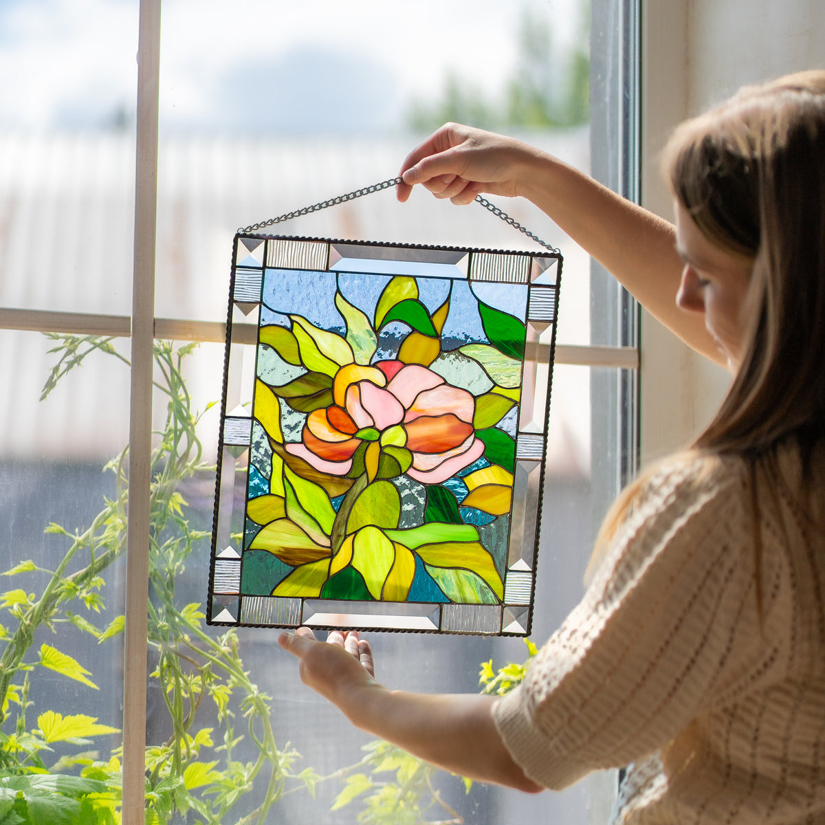 Woman holding a stained glass peony panel at her side, showing the pink flower and leaf design against a soft indoor background.