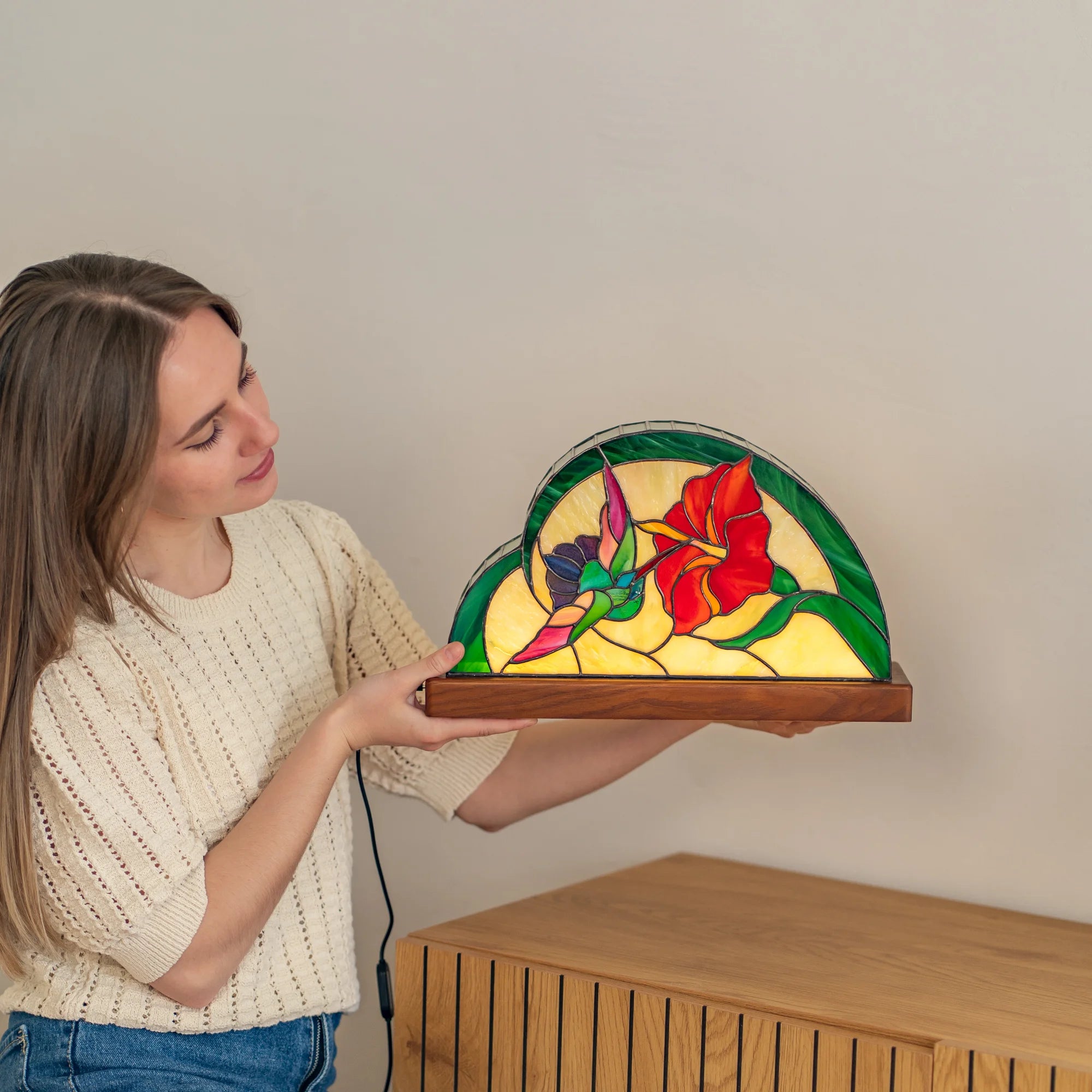 Woman holding a glowing hummingbird stained glass table lamp on its wooden base, red hibiscus and green leaves lit from within - handmade Tiffany-style home decor and gift