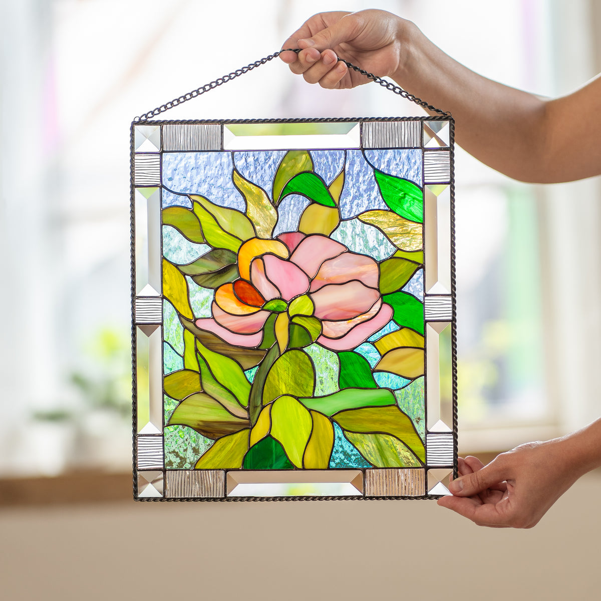Stained glass peony panel with beveled corners displayed upright on a wooden console beside dried wheat stems and a potted plant.