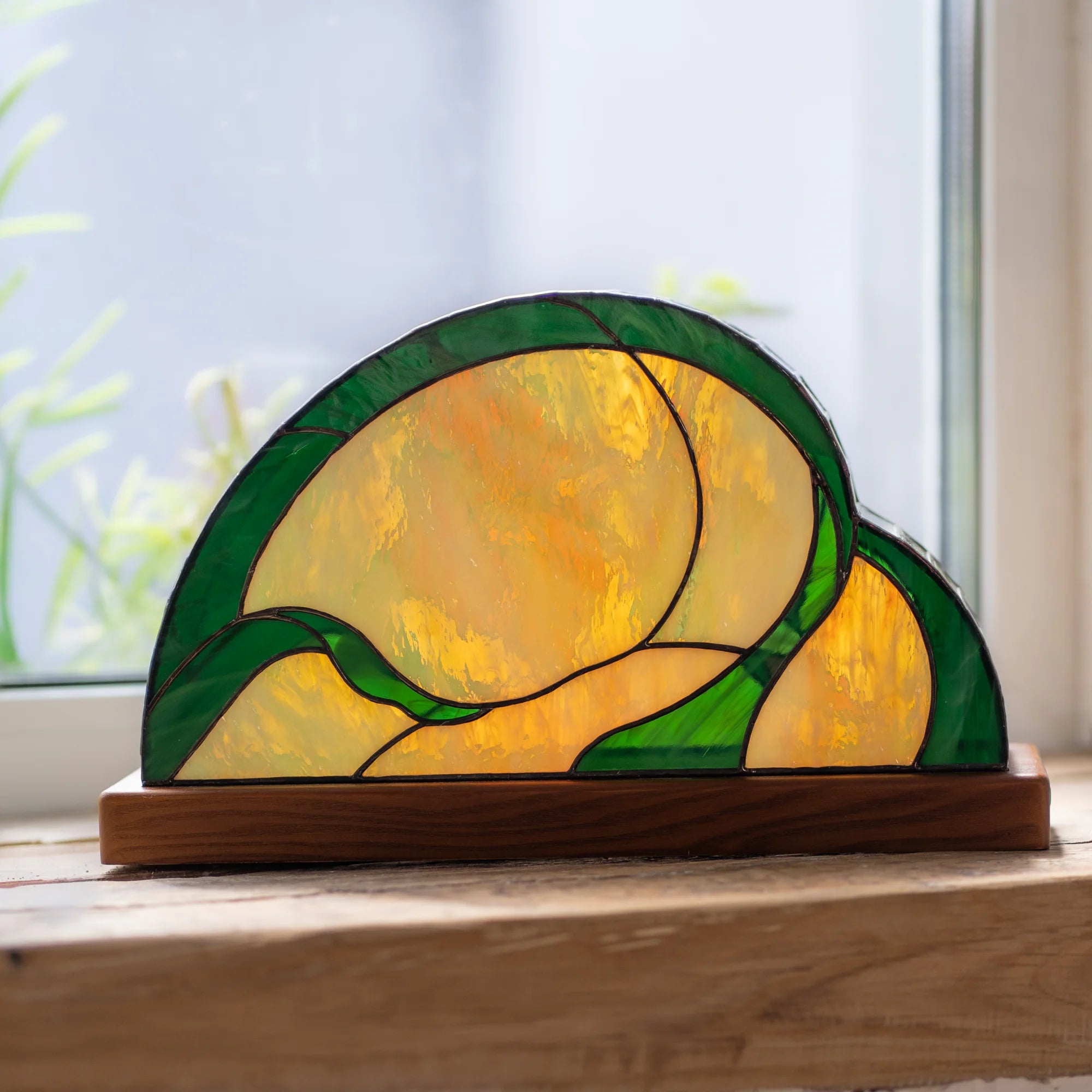 Back view of a hummingbird stained glass table lamp on a windowsill showing the amber and green glass and half-dome shape on its wooden base - handmade Tiffany-style lighting