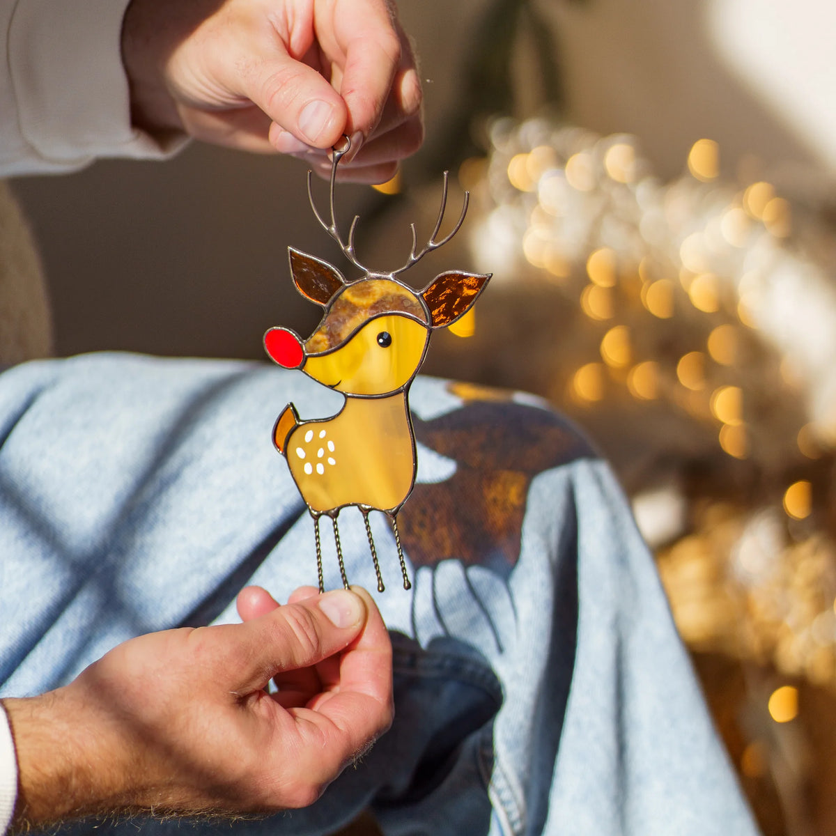 Man's hand holding the chain of a stained glass reindeer suncatcher with a red nose and antlers, warm string lights in the background.