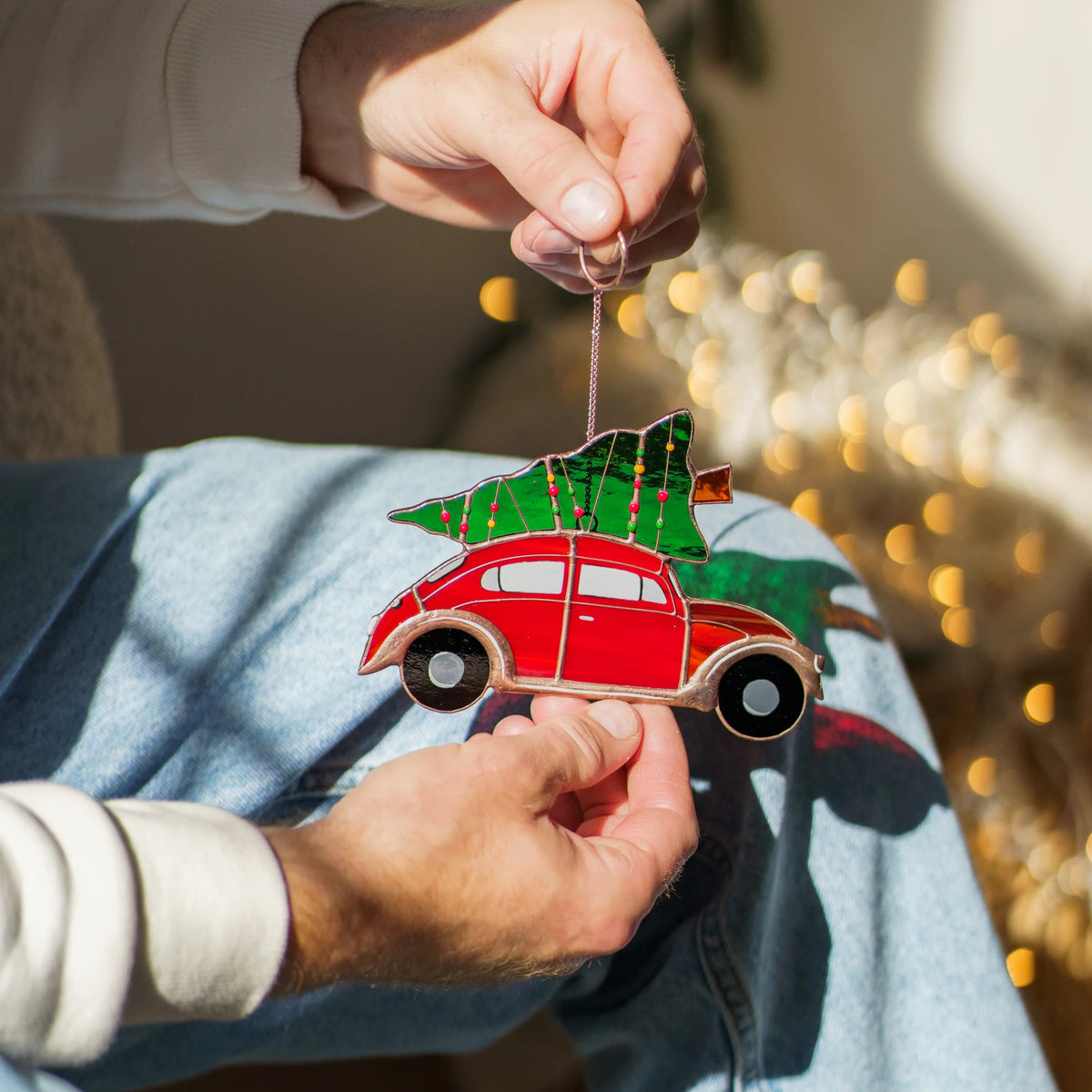 Man's hand holding the chain of a stained glass red car suncatcher topped with a Christmas tree, candy cane pillows in the background.