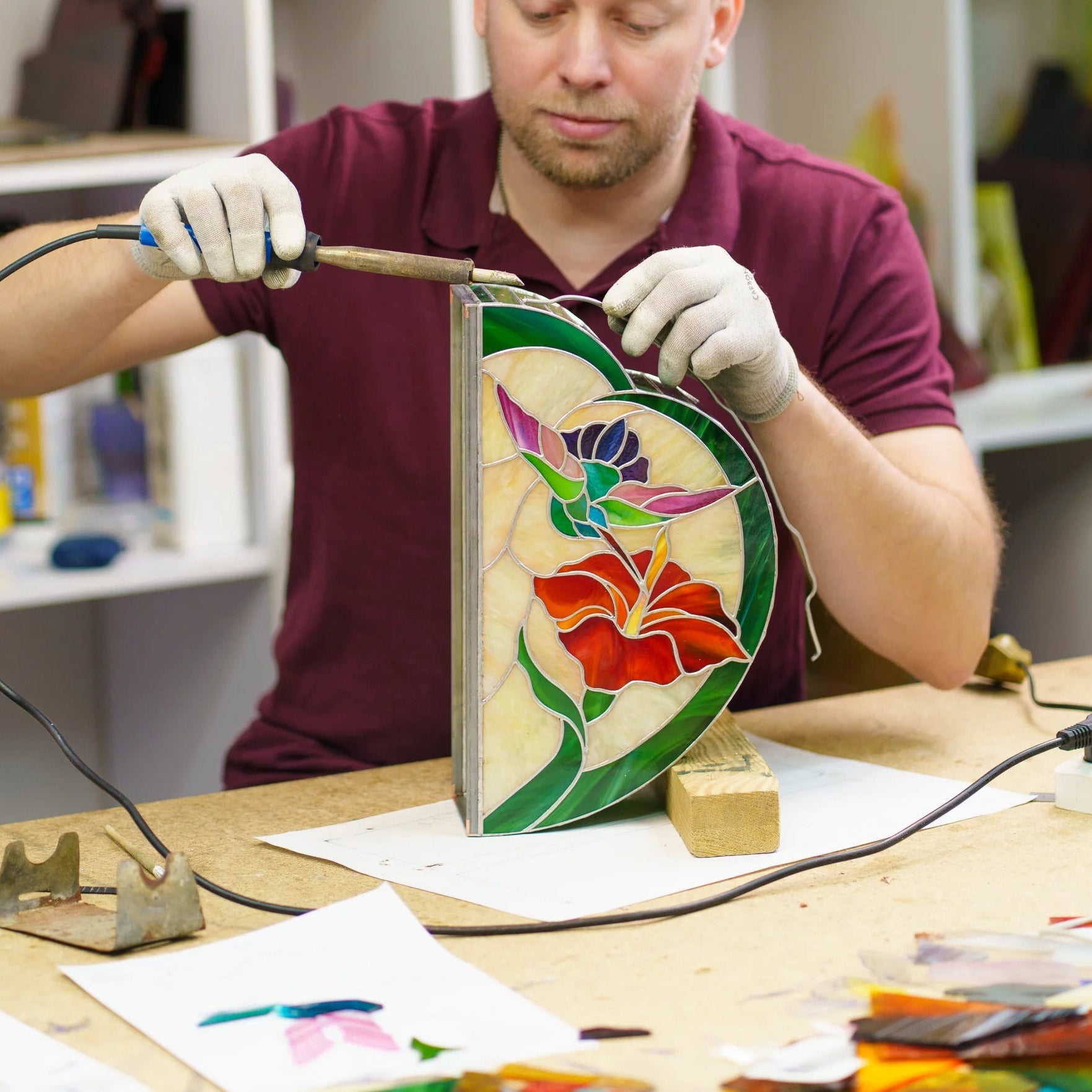 Glass artist soldering a hummingbird and hibiscus stained glass lamp in the studio, hand-cutting and foiling each piece - handmade Tiffany-technique craftsmanship since 1995
