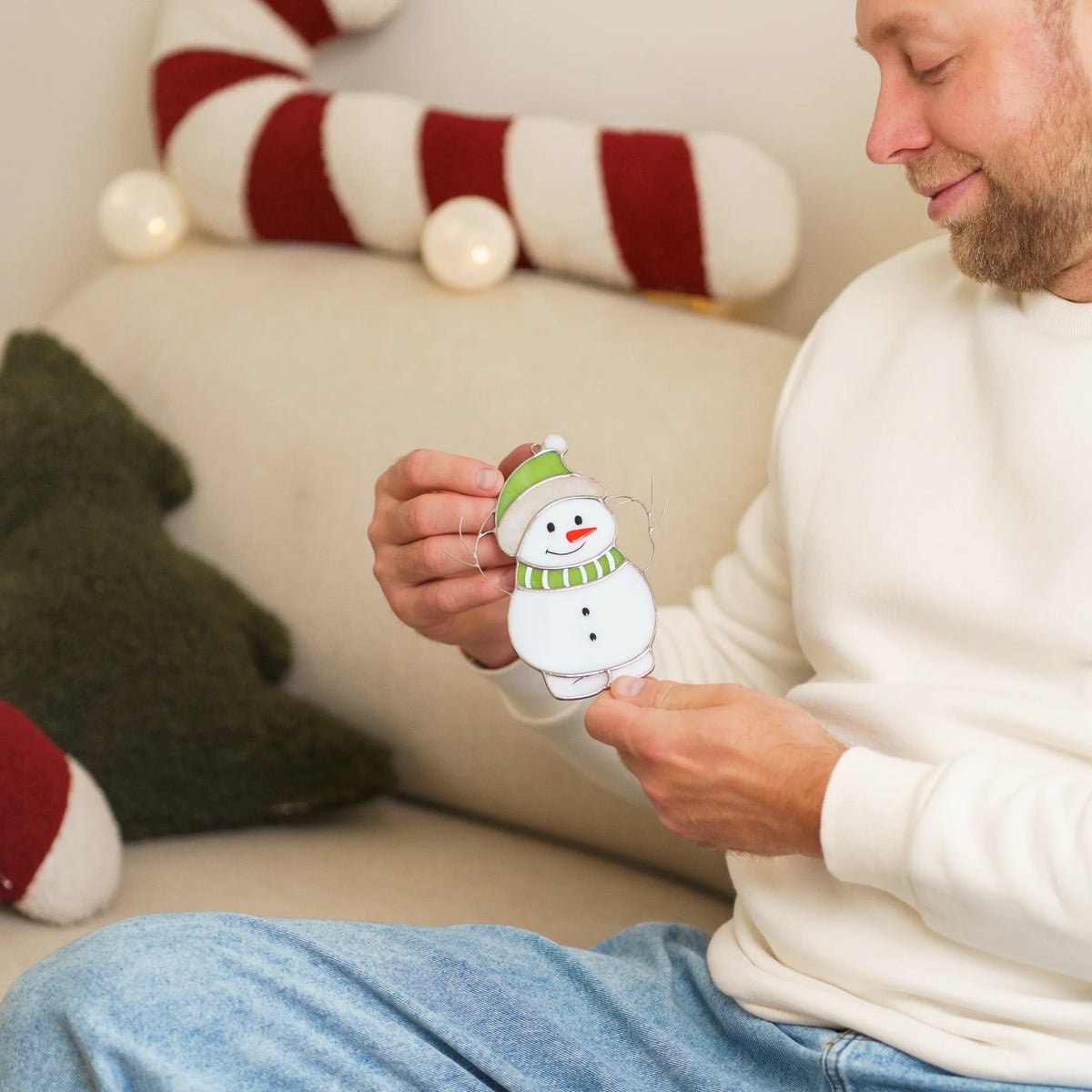 Man sitting among Christmas pillows, looking closely down at a stained glass snowman suncatcher wearing a green hat and scarf.