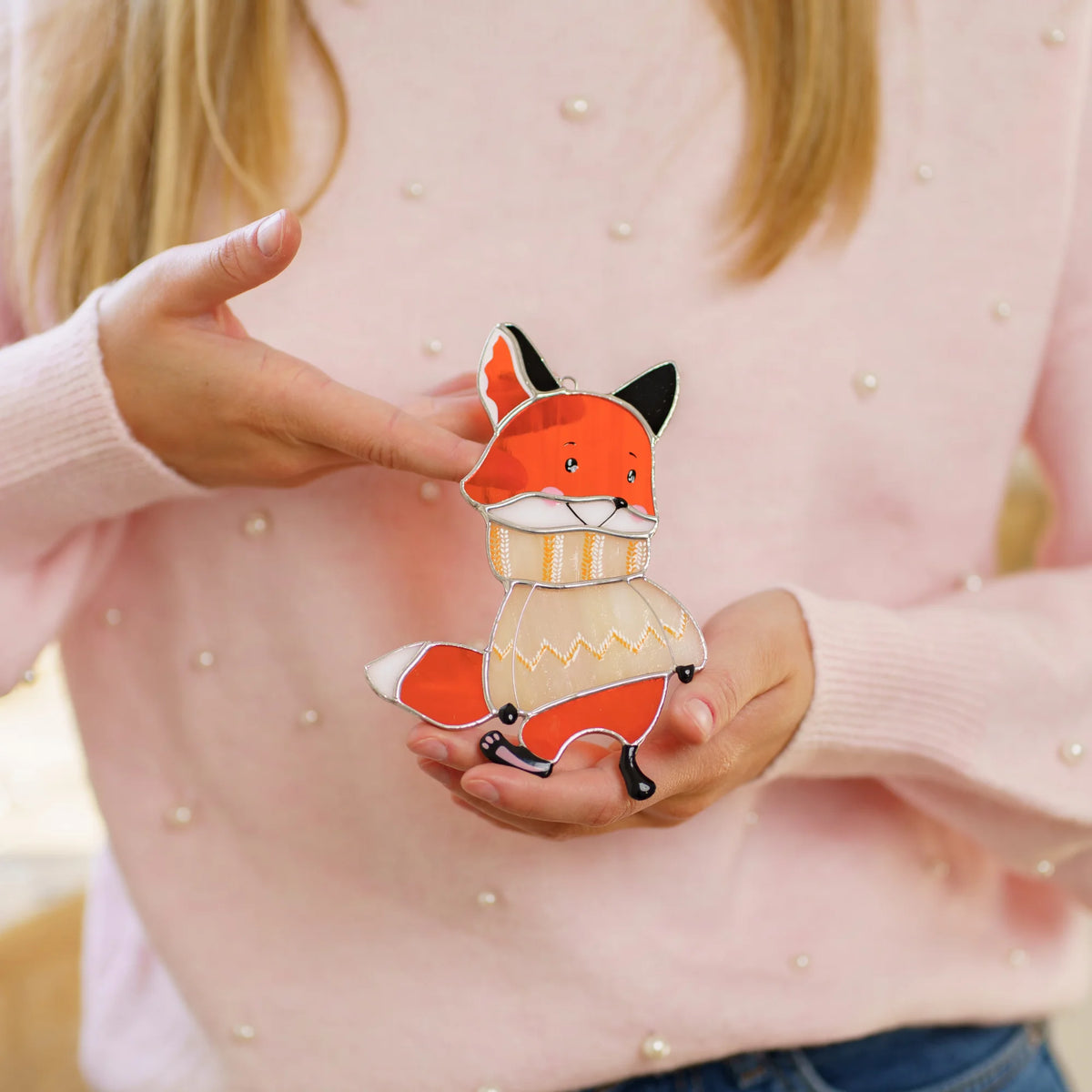 Girl in a pink sweater holding a handmade stained glass fox suncatcher wearing its own cozy knit sweater close in her hands.