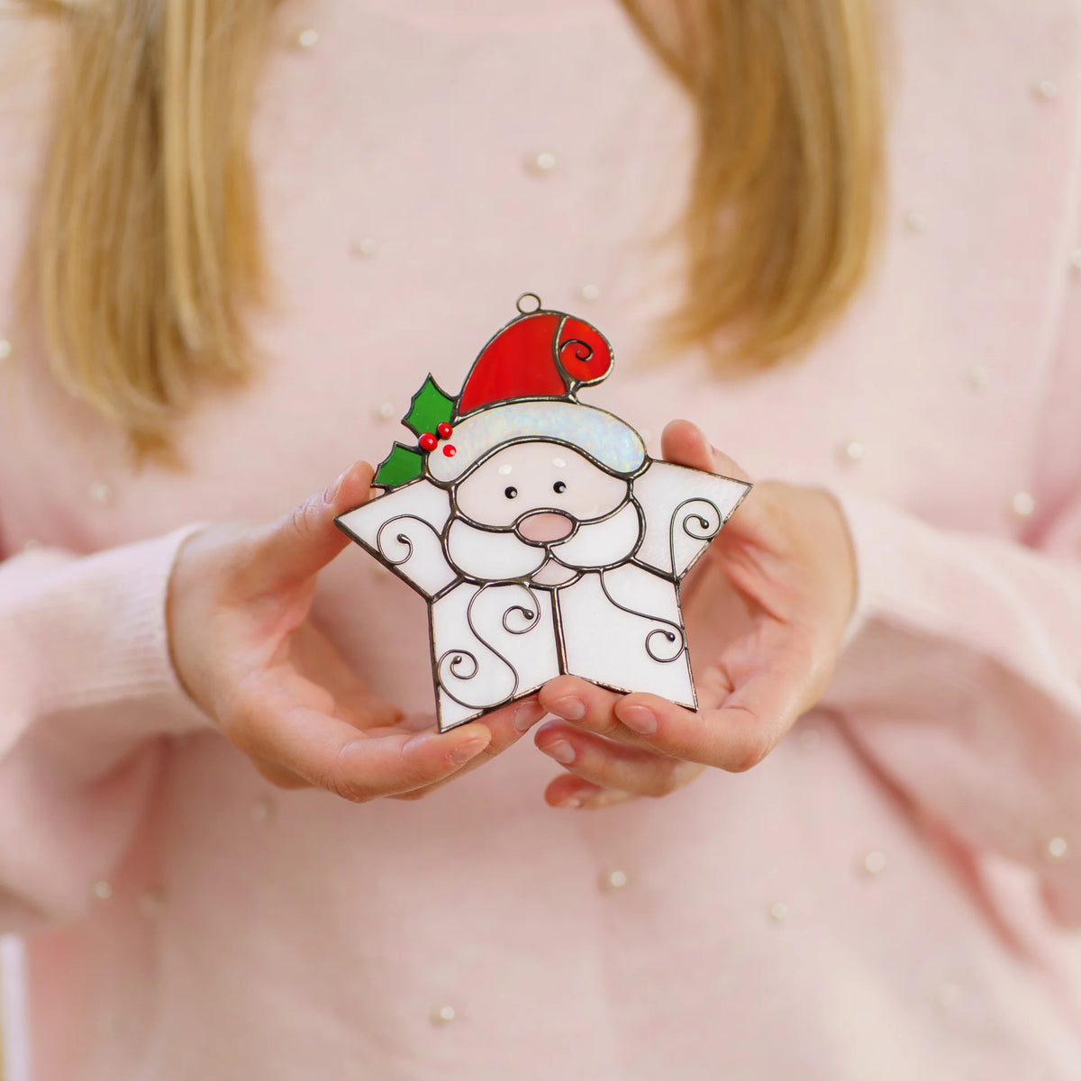 Woman in a pink sweater cradling a handmade Snowflake Santa stained glass suncatcher in a red hat with green holly and painted red berries - a sweet Christmas ornament and a cheerful holiday gift