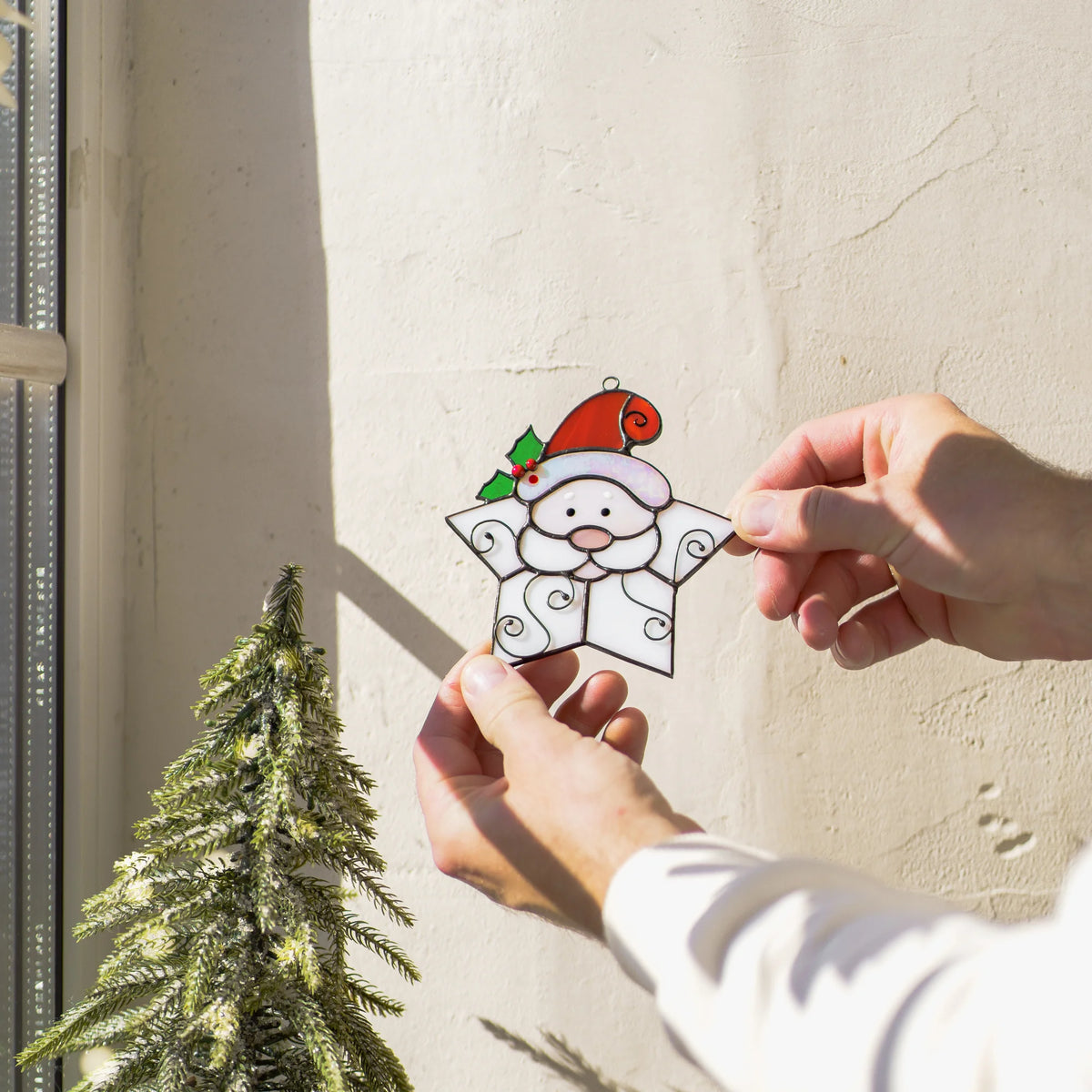 Snowflake Santa Christmas stained glass suncatcher held up to a sunlit wall beside a small frosted Christmas tree, its white star shape glowing softly - handmade glass Christmas decor for windows
