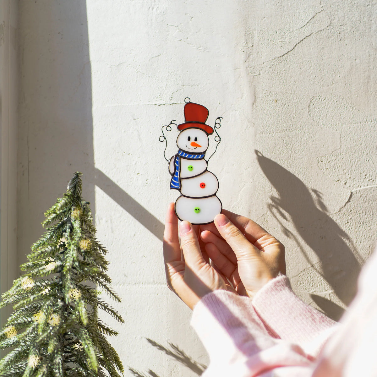 Christmas stained glass snowman held up to a sunlit wall beside a small frosted Christmas tree, its curly wire arms casting soft shadows - handmade glass Christmas decor for your windows