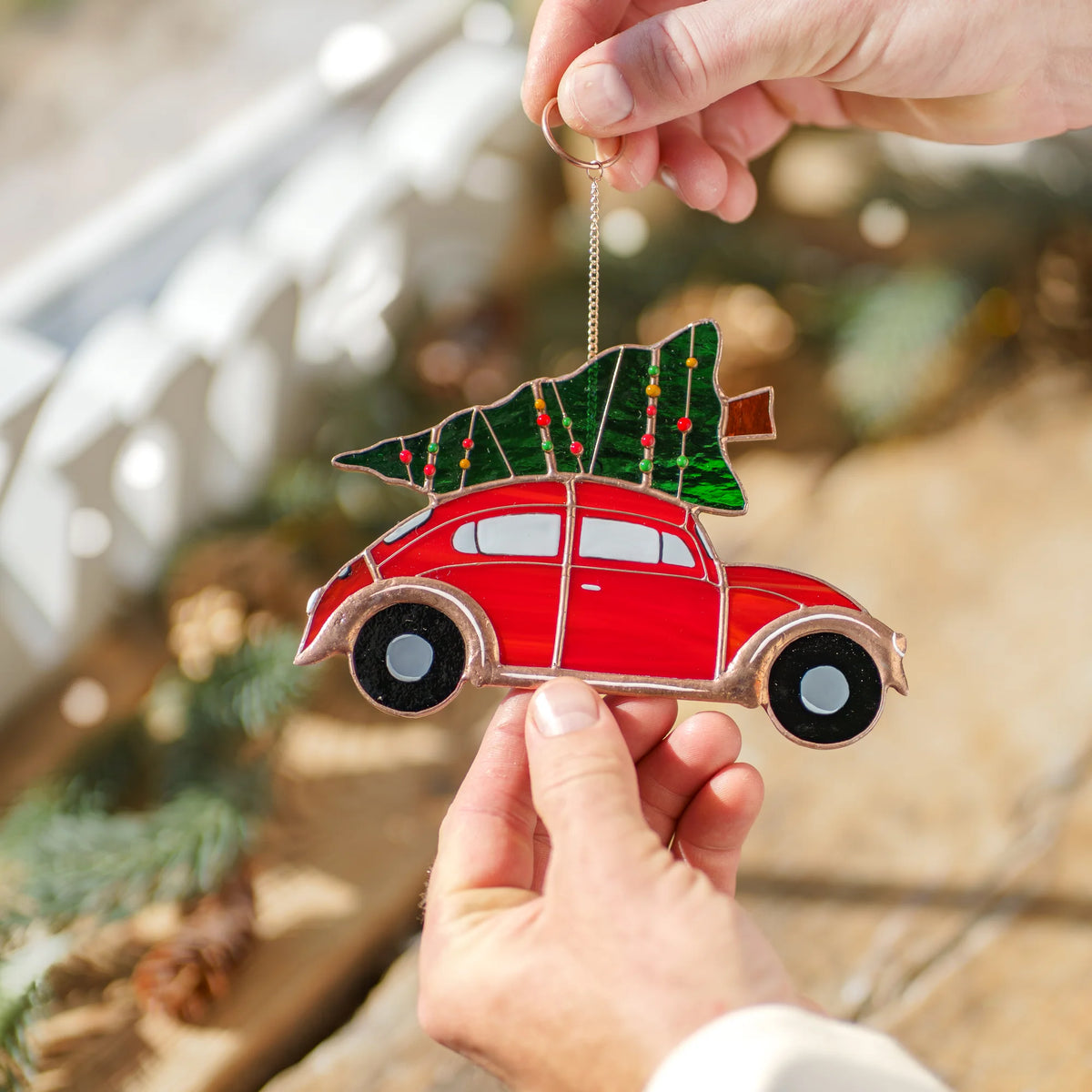 Two hands holding a stained glass red car suncatcher with a Christmas tree on its roof outdoors near pine branches and stone.