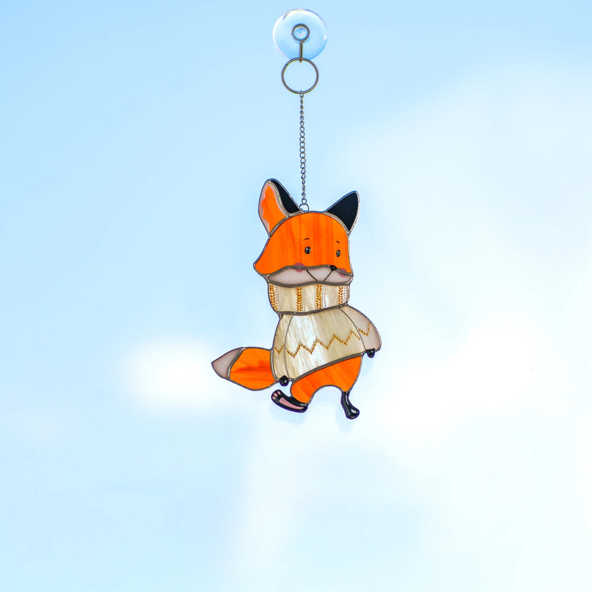 Hand holding the chain of a stained glass fox suncatcher in a sweater against a sunlit white wall with a Christmas tree branch nearby.