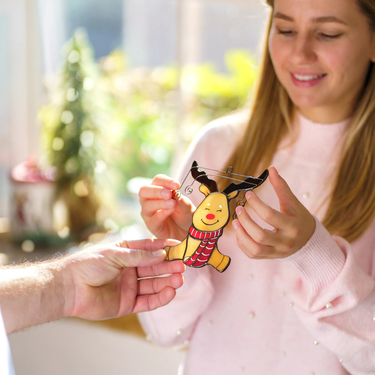 A person in a light pink sweater holds a Christmas staiend glass ornament shaped like Rudolph the Red-Nosed Reindeer, with another hand holding a metal tool near it, against a softly blurred indoor background.