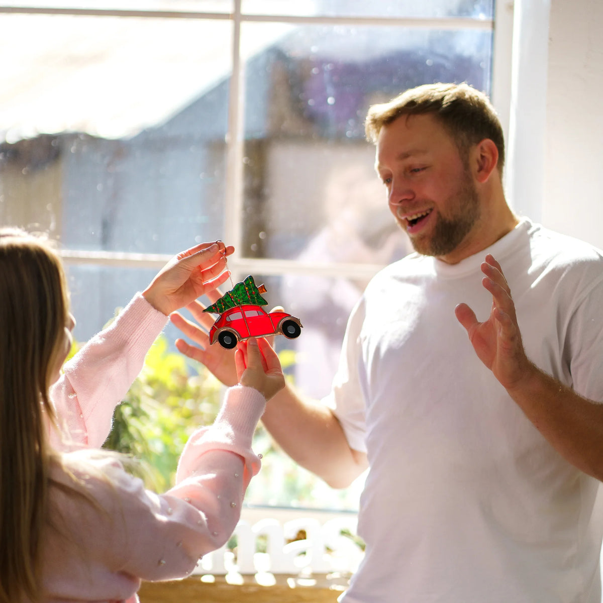 Man holding up a stained glass red car suncatcher to show a smiling woman reaching for it, sunlight streaming through a window.
