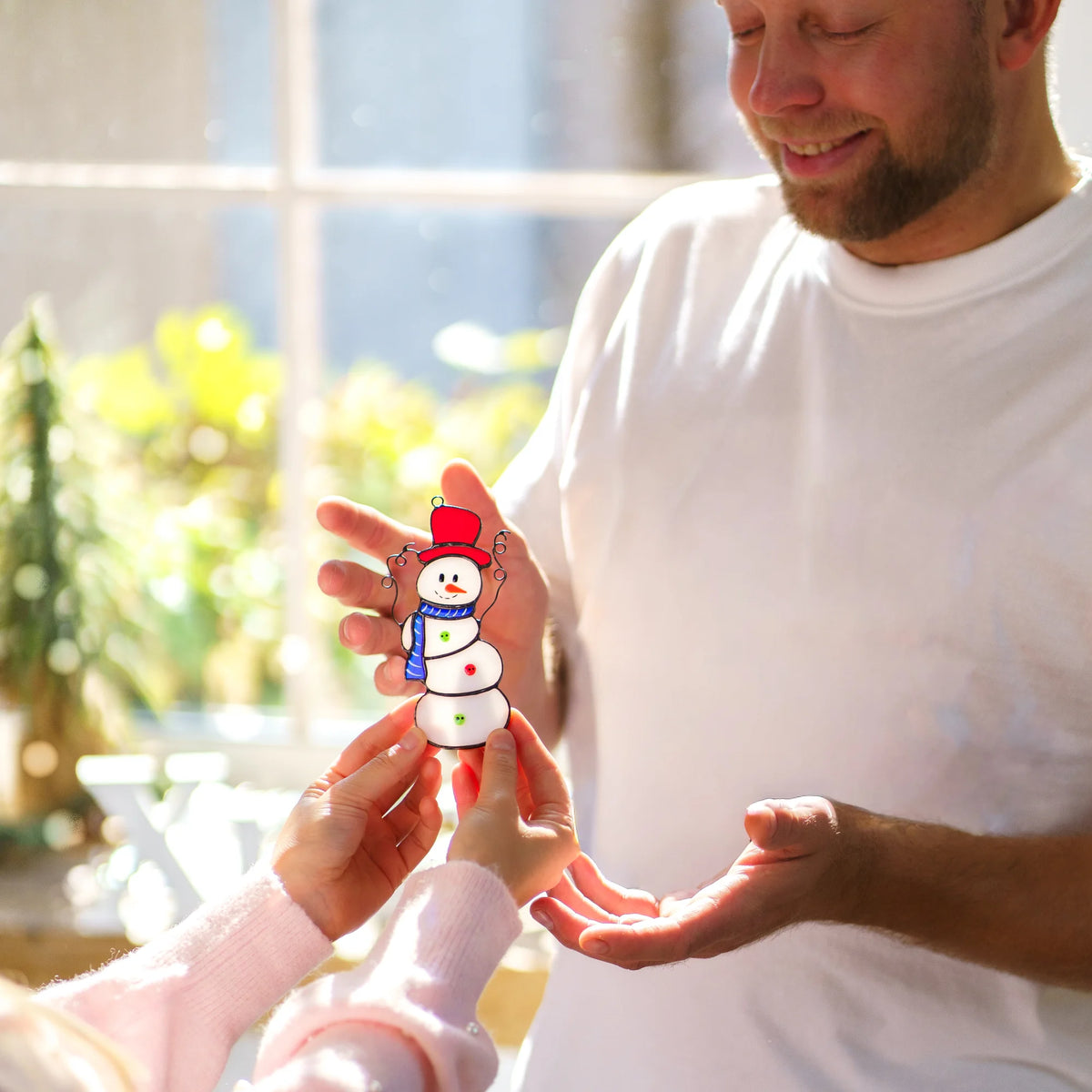 Small hands passing a handmade Christmas stained glass snowman in a red top hat and blue scarf to a smiling man by a sunny window - a heartwarming holiday gift and colorful Christmas suncatcher